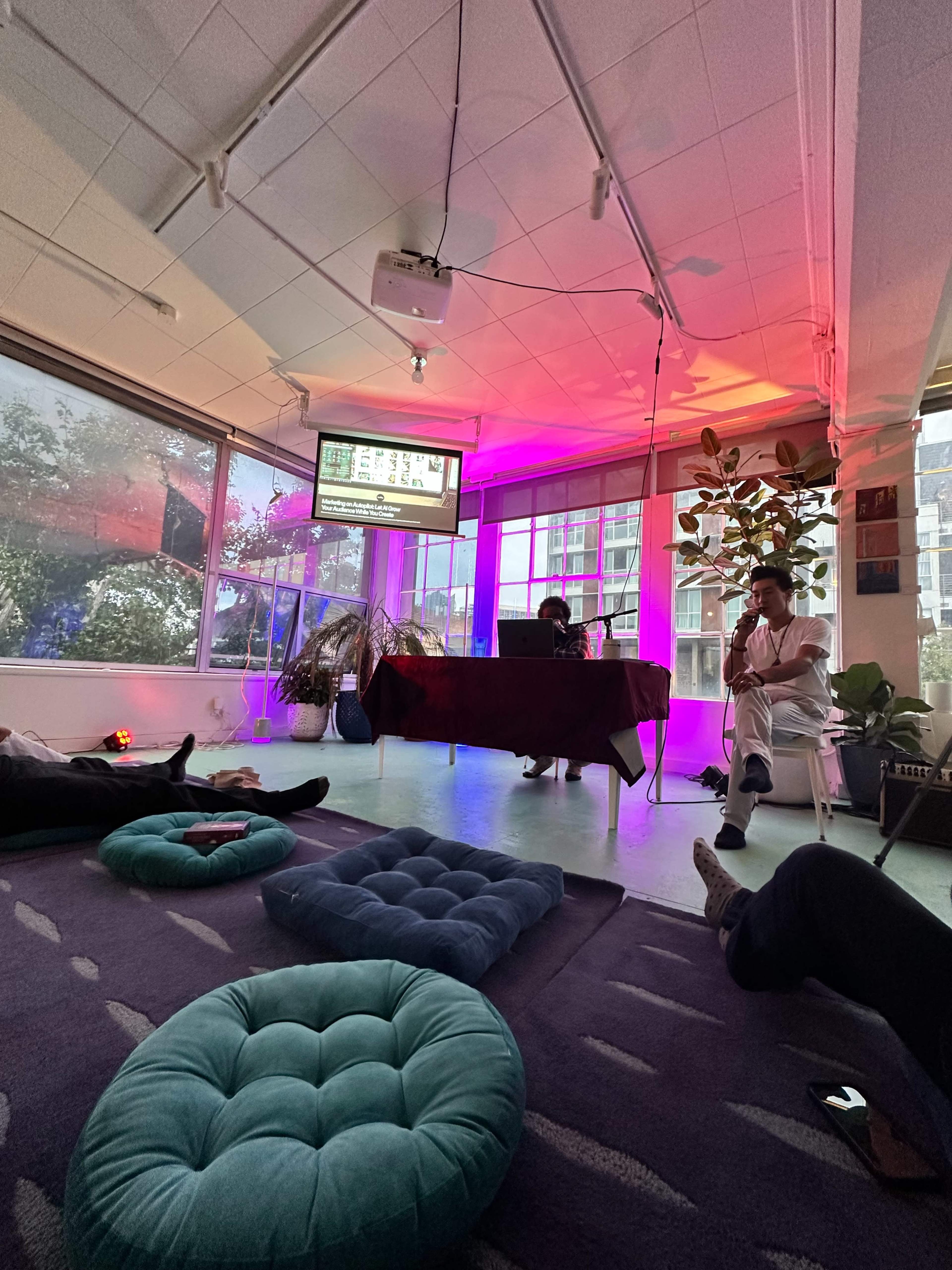 The image shows a brightly lit indoor space with colorful lighting, where two people sit at a table in front of a screen, while others recline on cushions scattered on the floor.