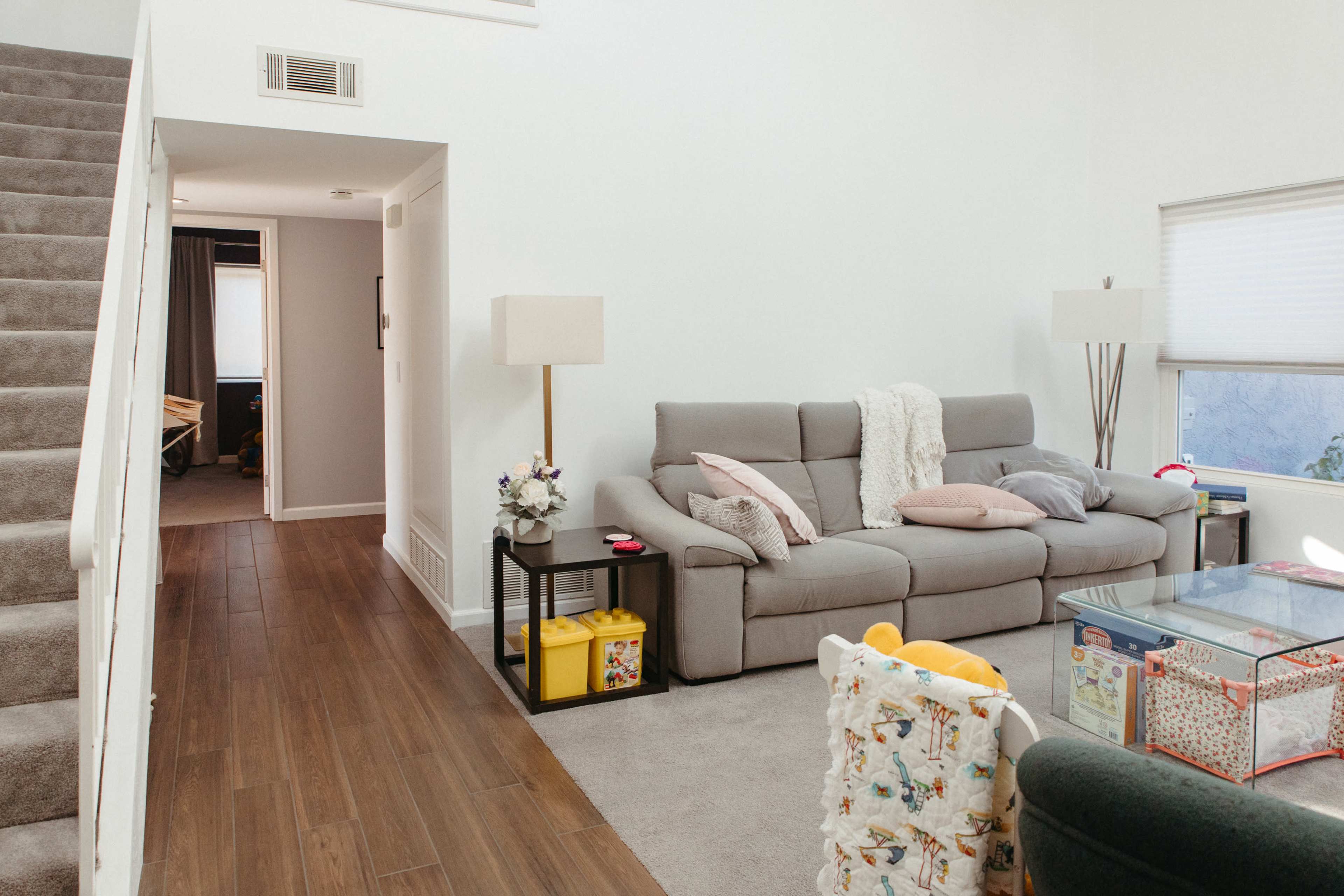 The image shows a living room with a gray sofa, a coffee table, and a staircase leading to the upper level.