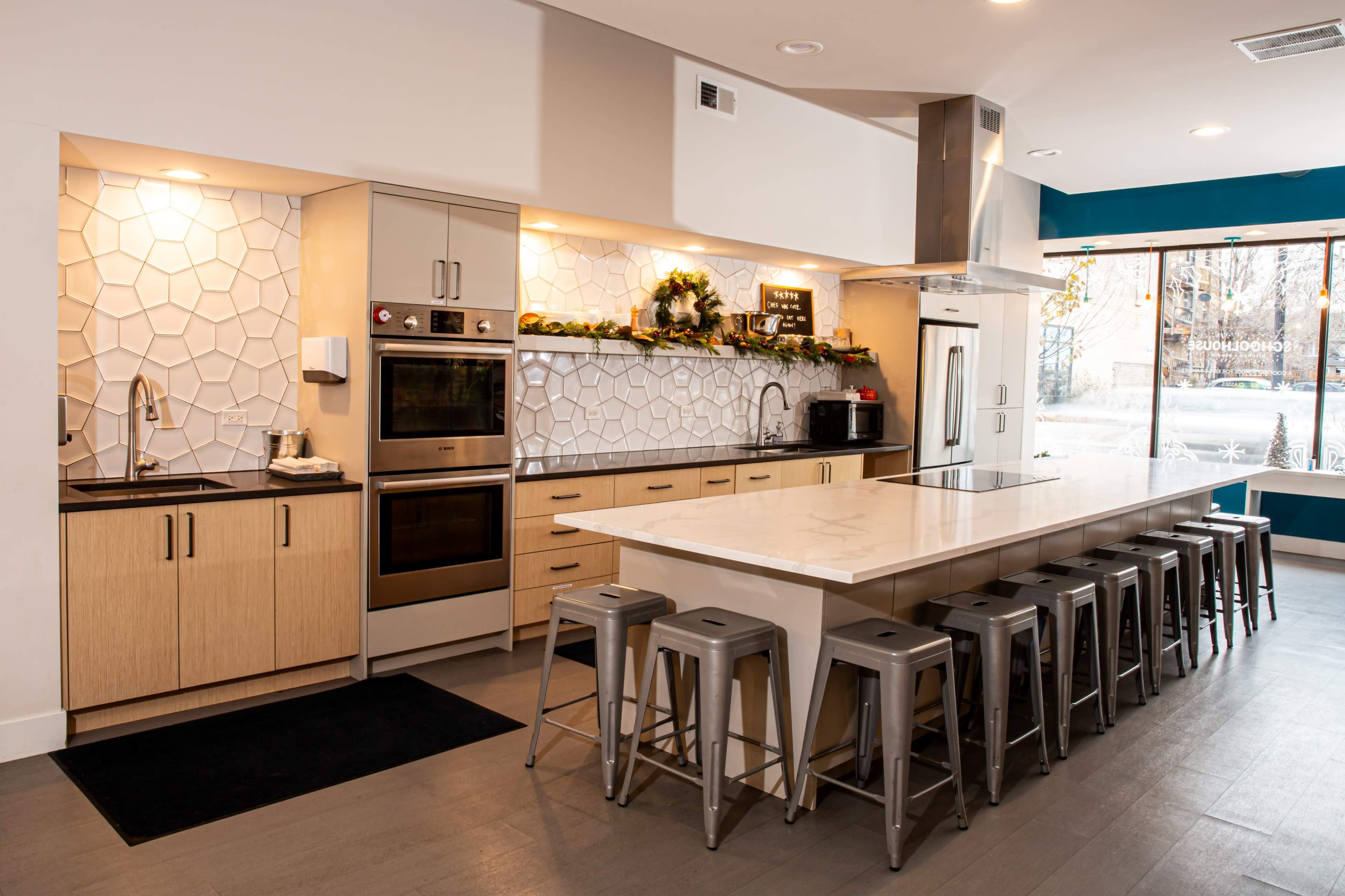 A modern kitchen with a long island counter, metal stools, and geometric white tile backsplash.