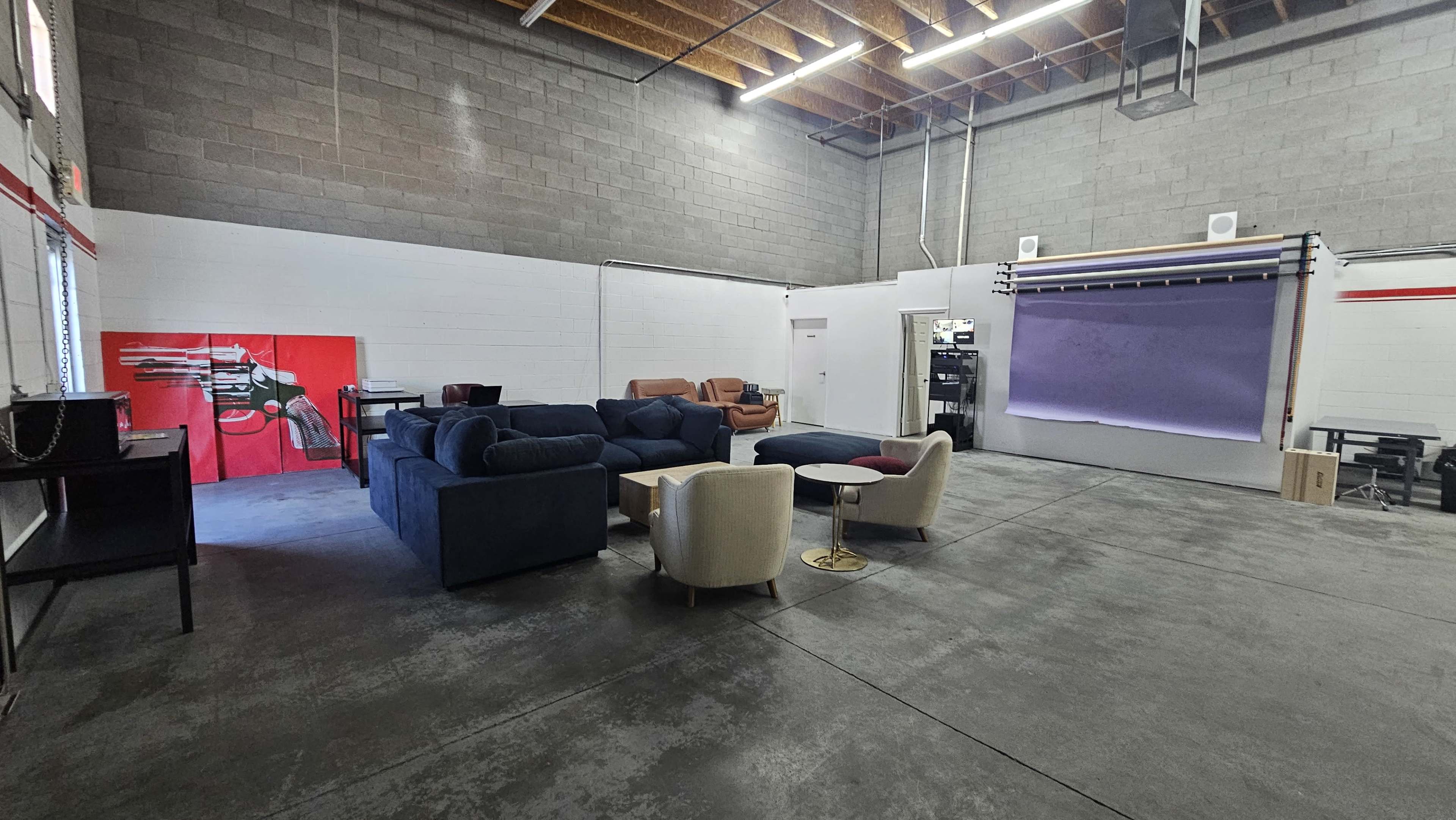 The image shows an industrial-style interior space with a seating area featuring various sofas and chairs, and a wall adorned with a red artwork.