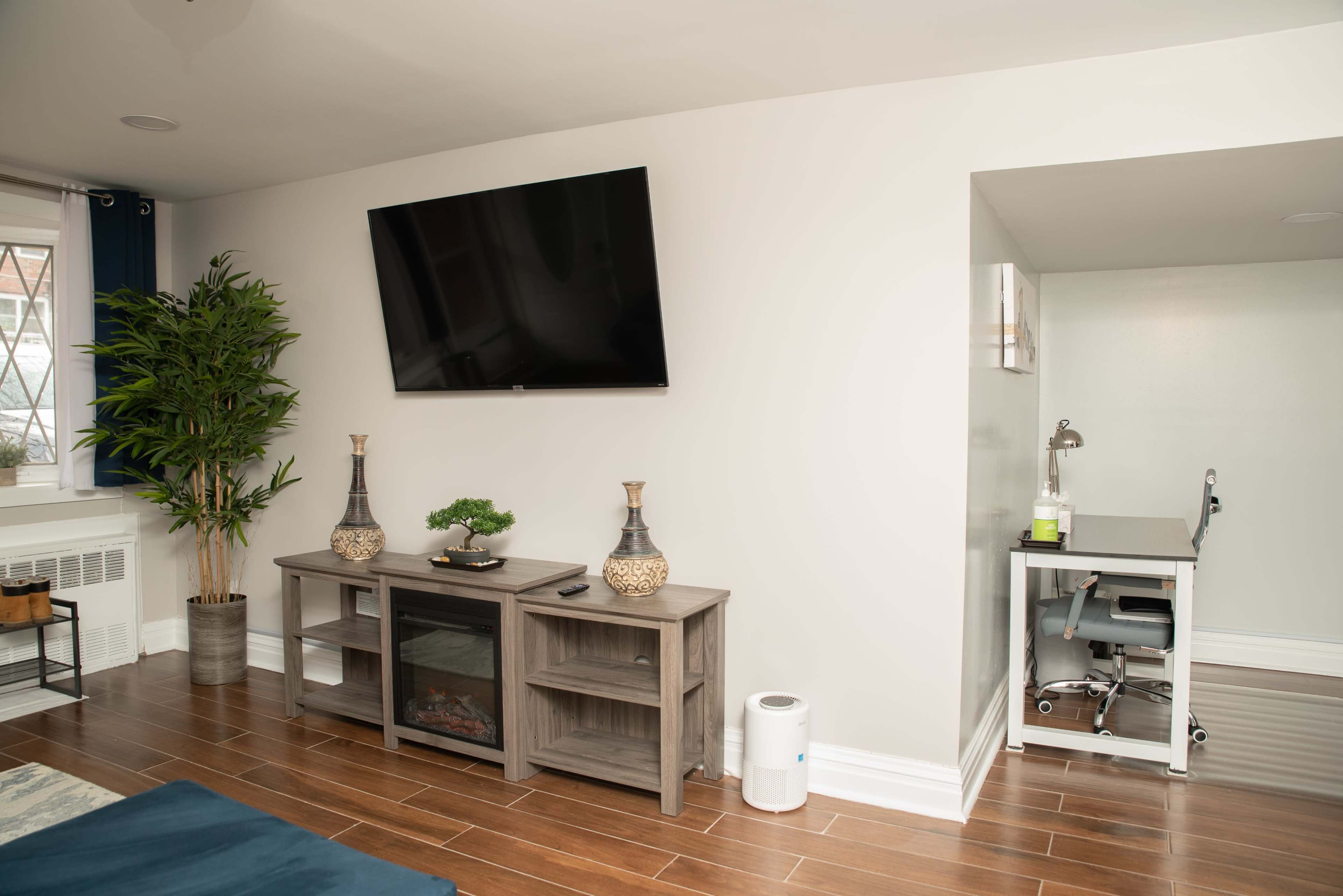 A living room features a television mounted on the wall, a small table with chairs, and a light-colored sofa near a window.