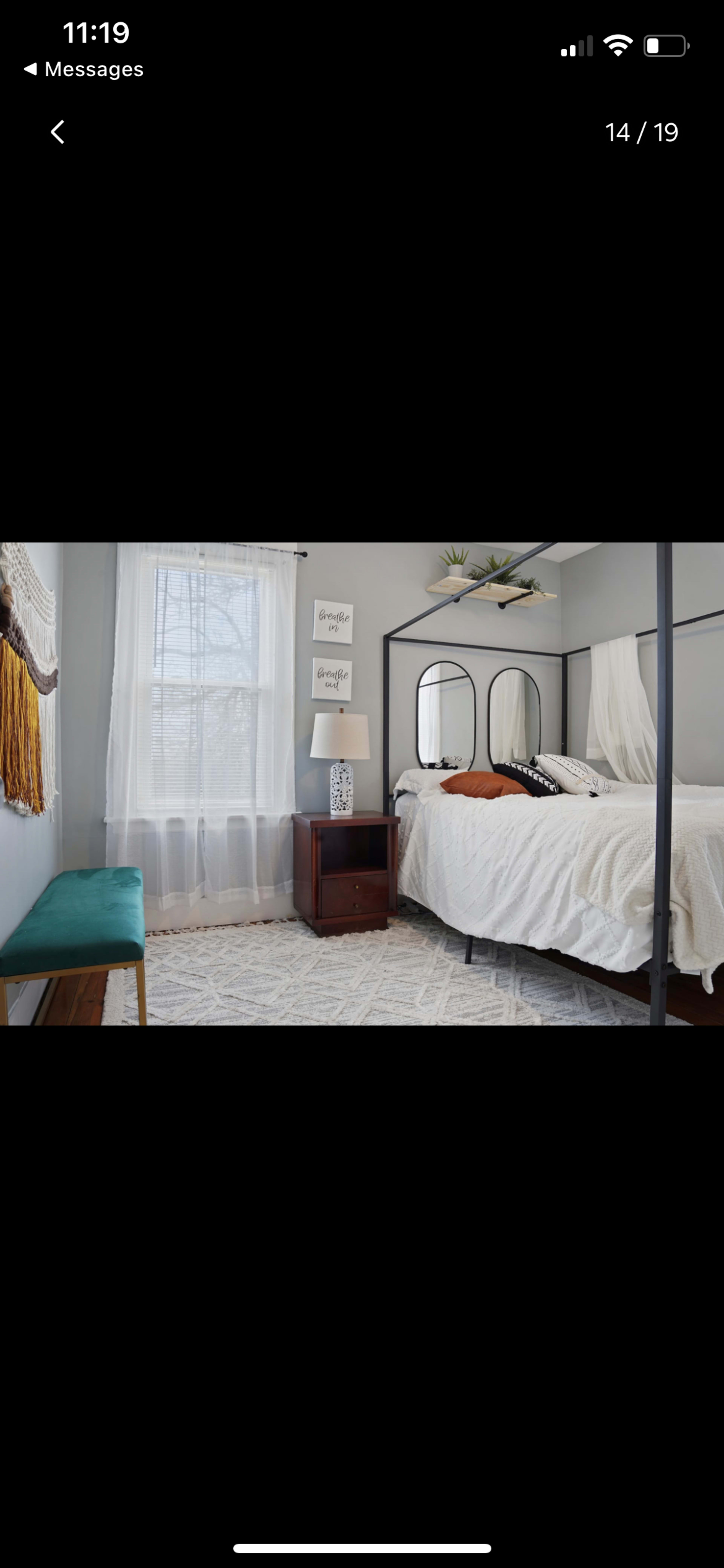 The image shows a bedroom with a bed, a nightstand, a lamp, a decorative mirror, and a bench near a window with sheer curtains.