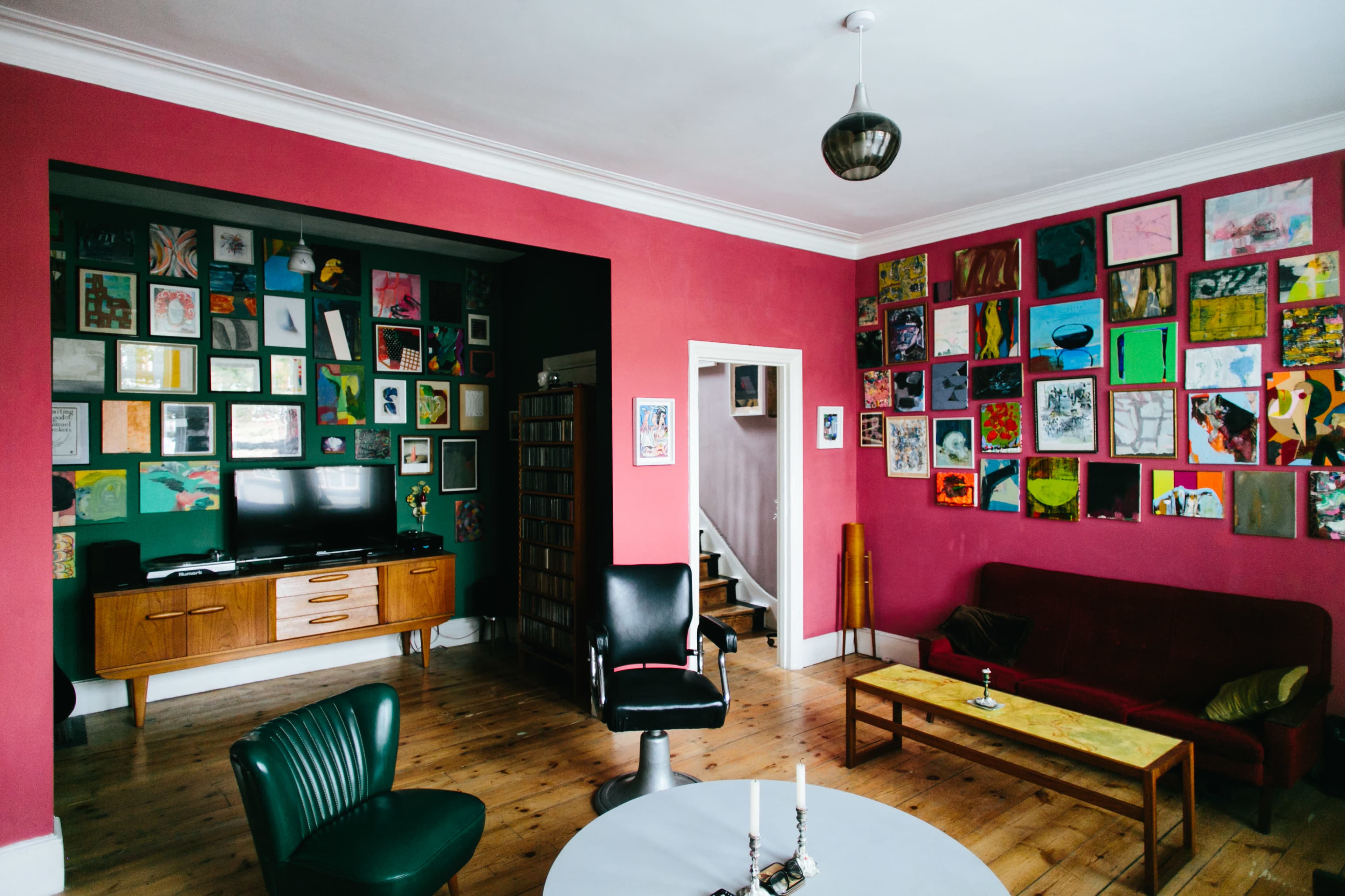 The image shows a living room with a red accent wall adorned with various colorful artworks, a wooden shelf with a television, and a combination of seating options including a dark red sofa and a green chair.