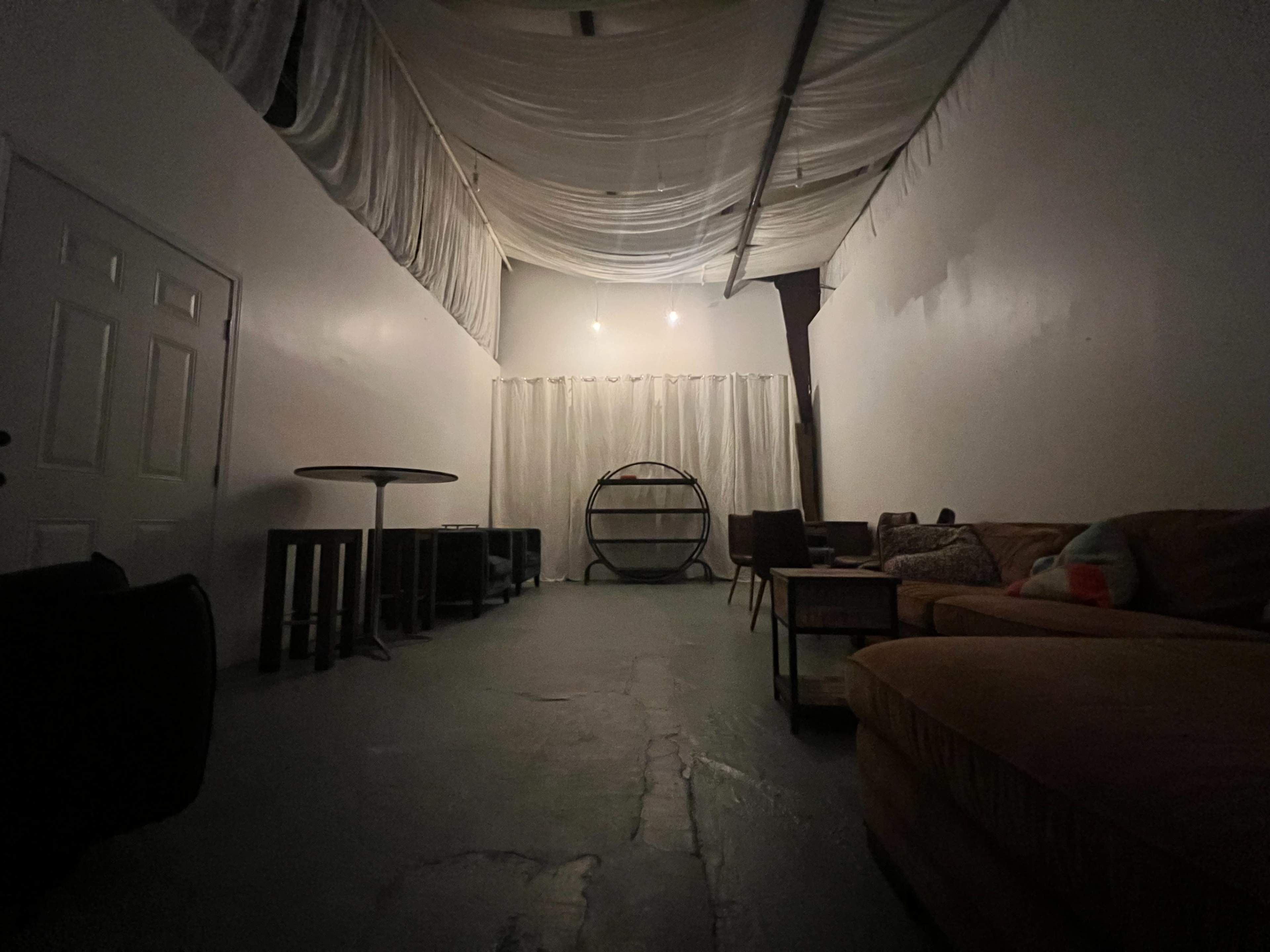 The image shows a dimly lit indoor space with a long ceiling draped in fabric, a circular shelving unit at the back, and a mix of furniture, including a couch and chairs, arranged throughout.