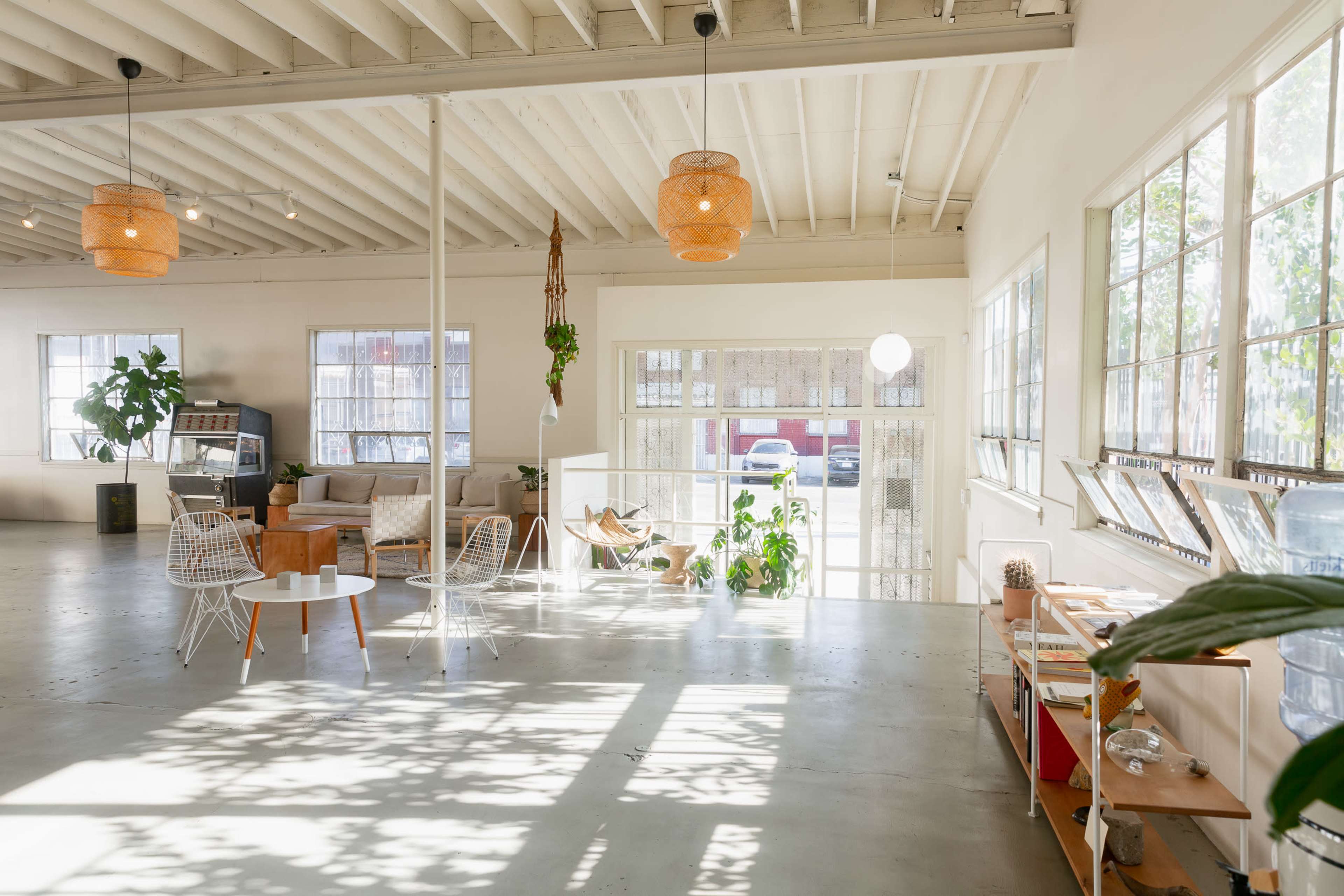 The image shows a spacious, well-lit interior featuring a seating area with modern furniture, large windows, and plants.