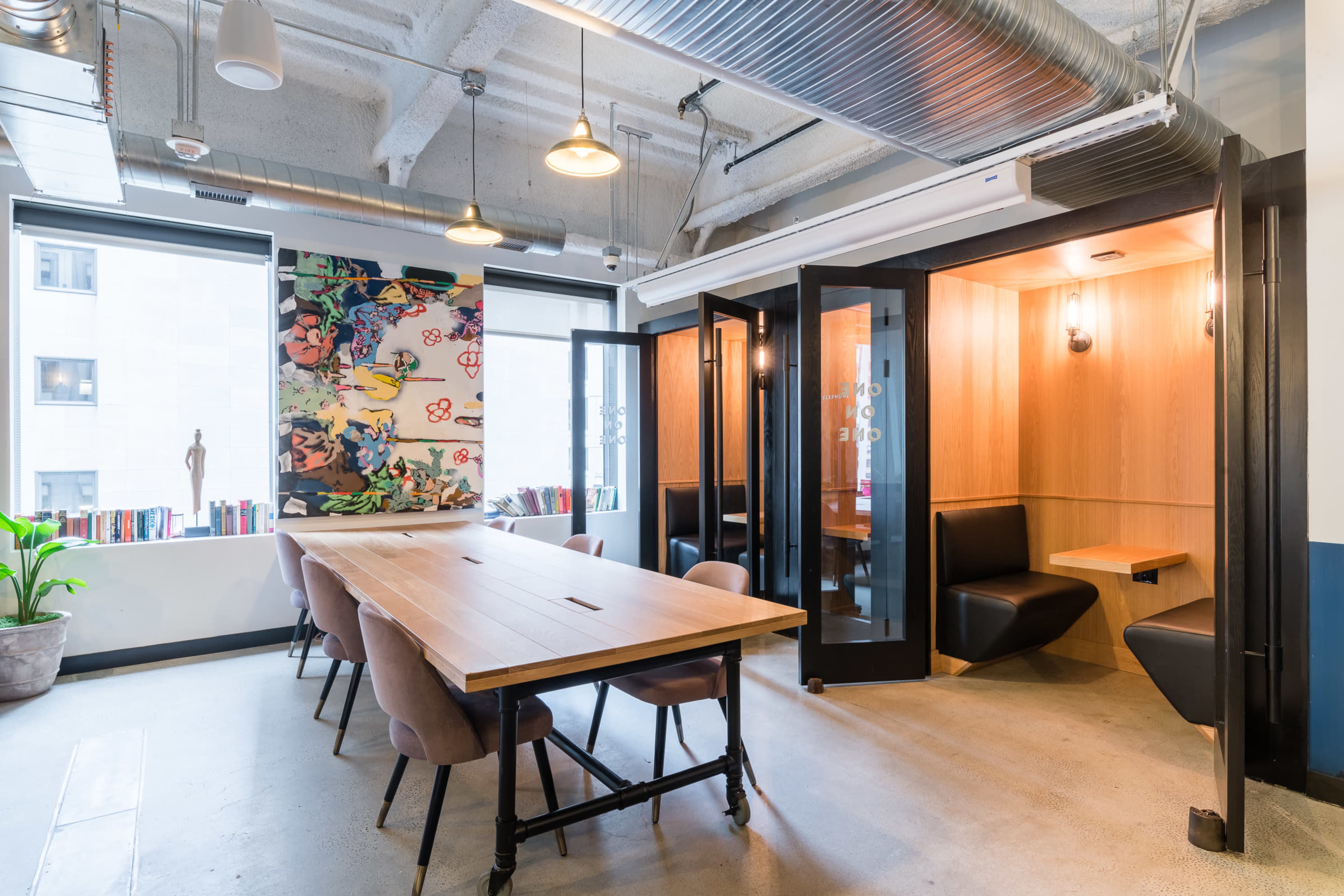 The image shows a modern meeting room with a long wooden table surrounded by chairs, and two enclosed booths along one side, decorated with artwork and plants throughout the space.