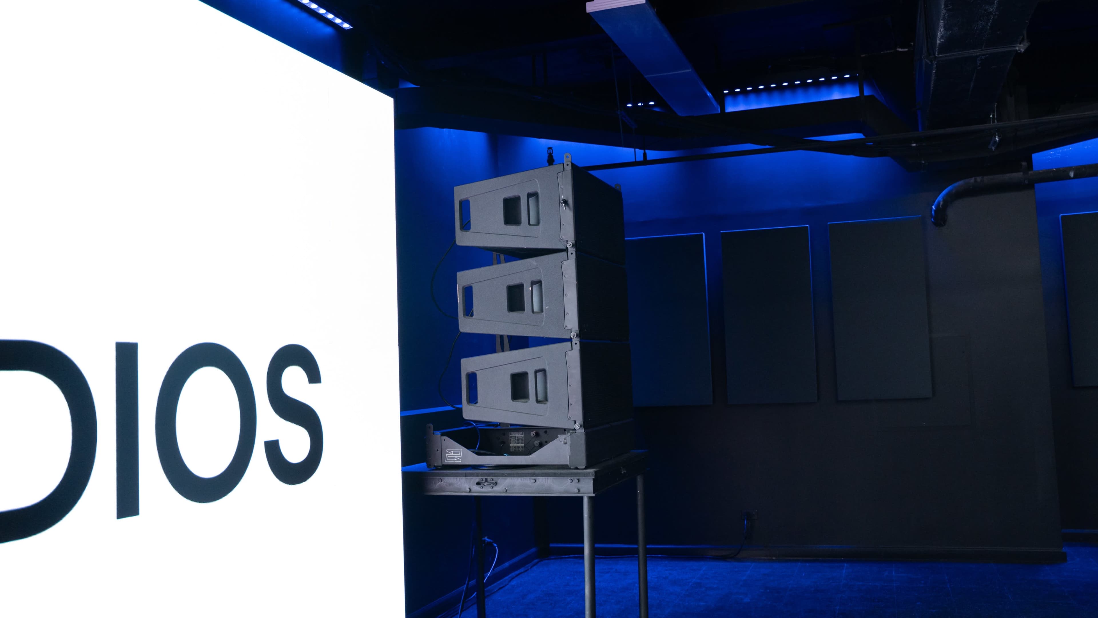 The image shows a stack of audio equipment placed on a table in a dimly lit room with blue ambient lighting and a large screen displaying the word "STUDIOS."