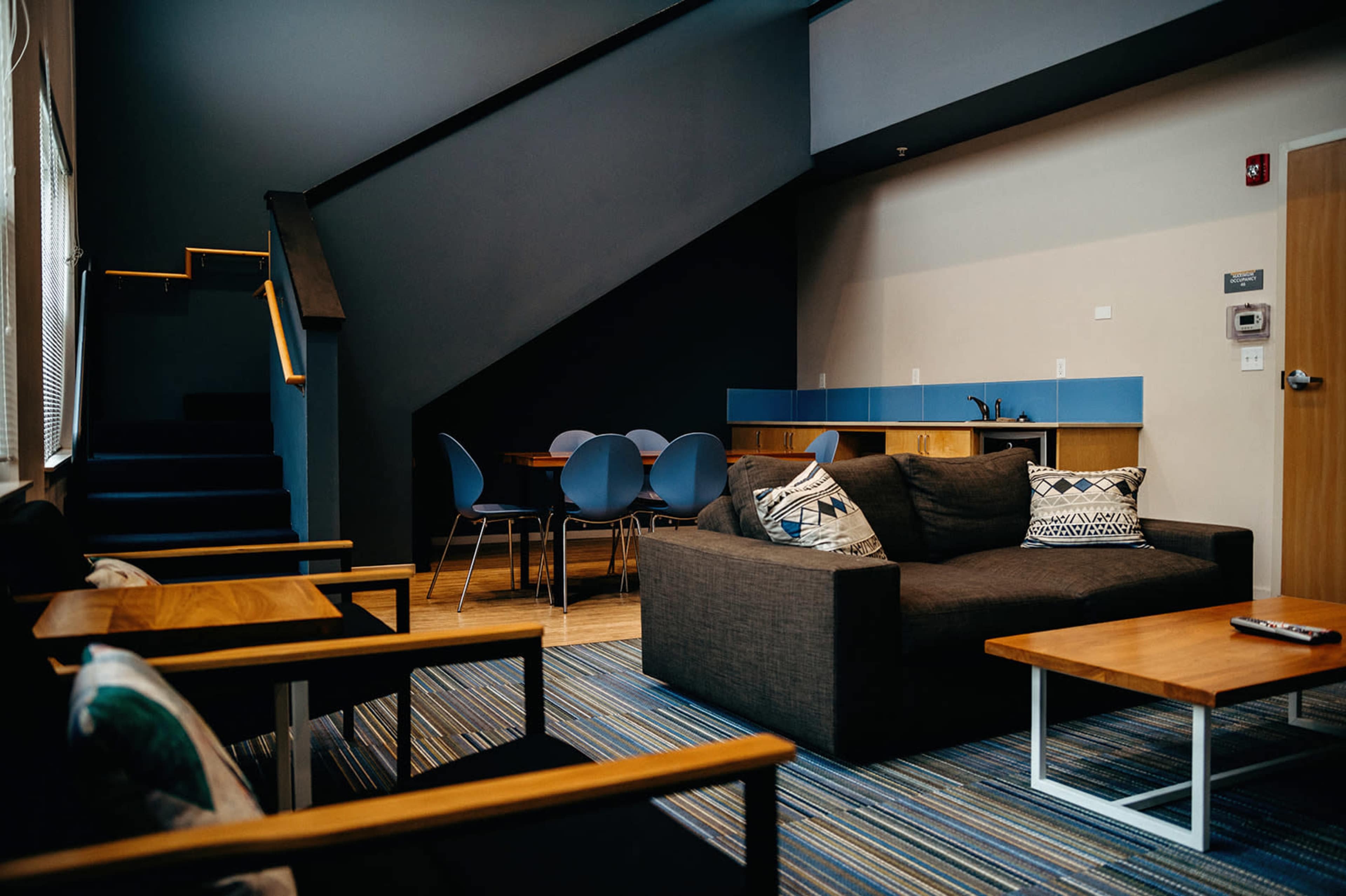 A modern lounge area features a brown sofa, wooden coffee tables, and a dining space with blue chairs adjacent to a staircase.