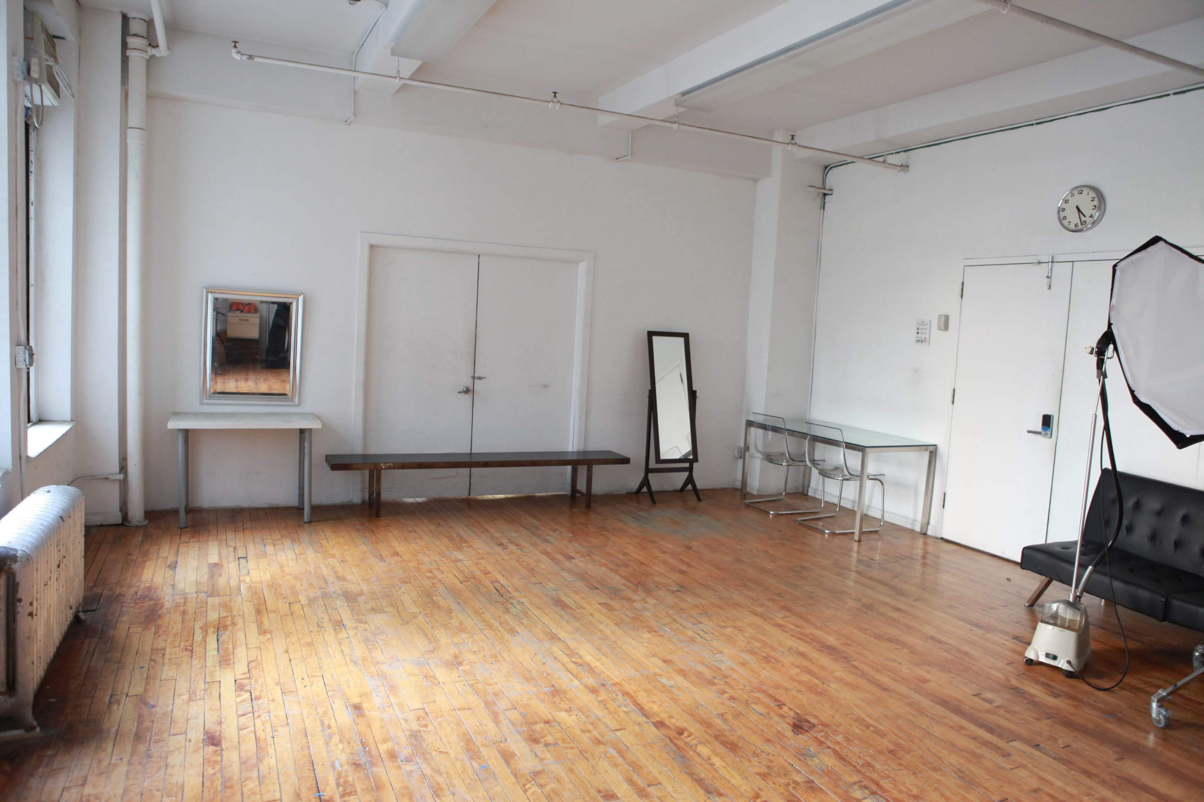 The image shows a spacious room with hardwood floors, featuring a bench, a couple of mirrors, a clock on the wall, a small table with chairs, and a black couch.