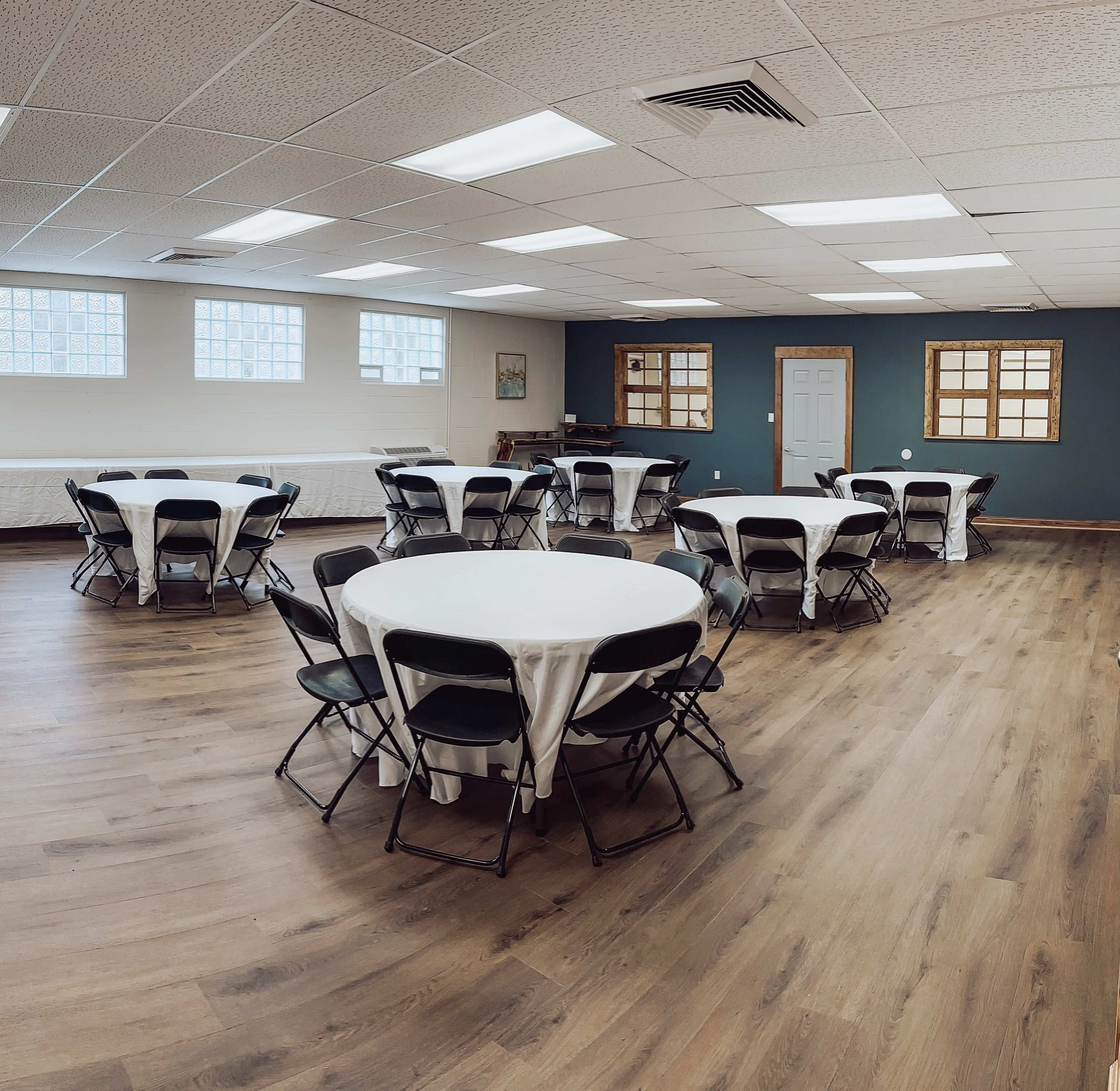 A spacious event room with several circular tables set with white tablecloths and black chairs arranged throughout.