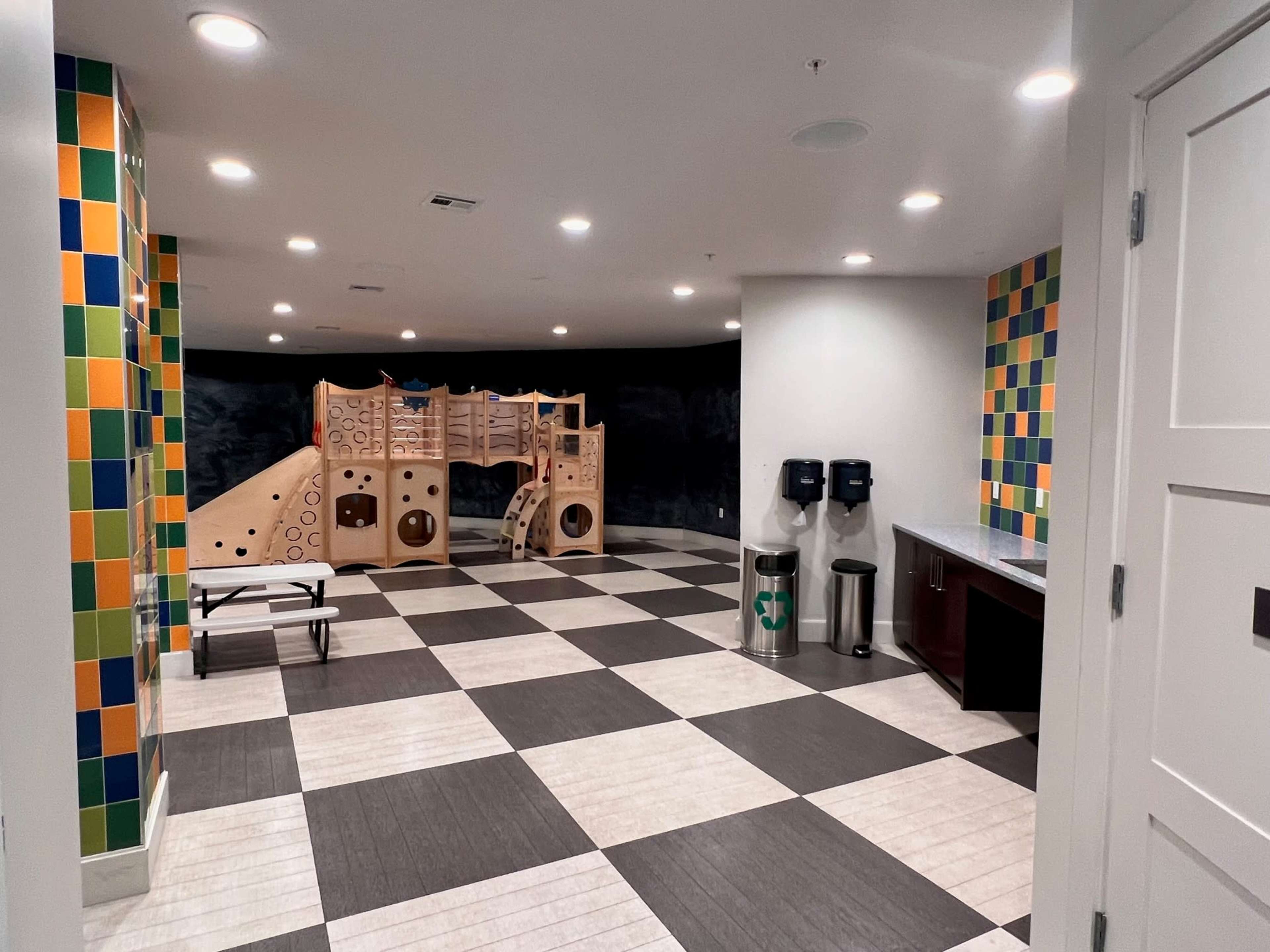 A brightly colored indoor play area featuring a wooden climbing structure, with tiled walls and a checkered floor.