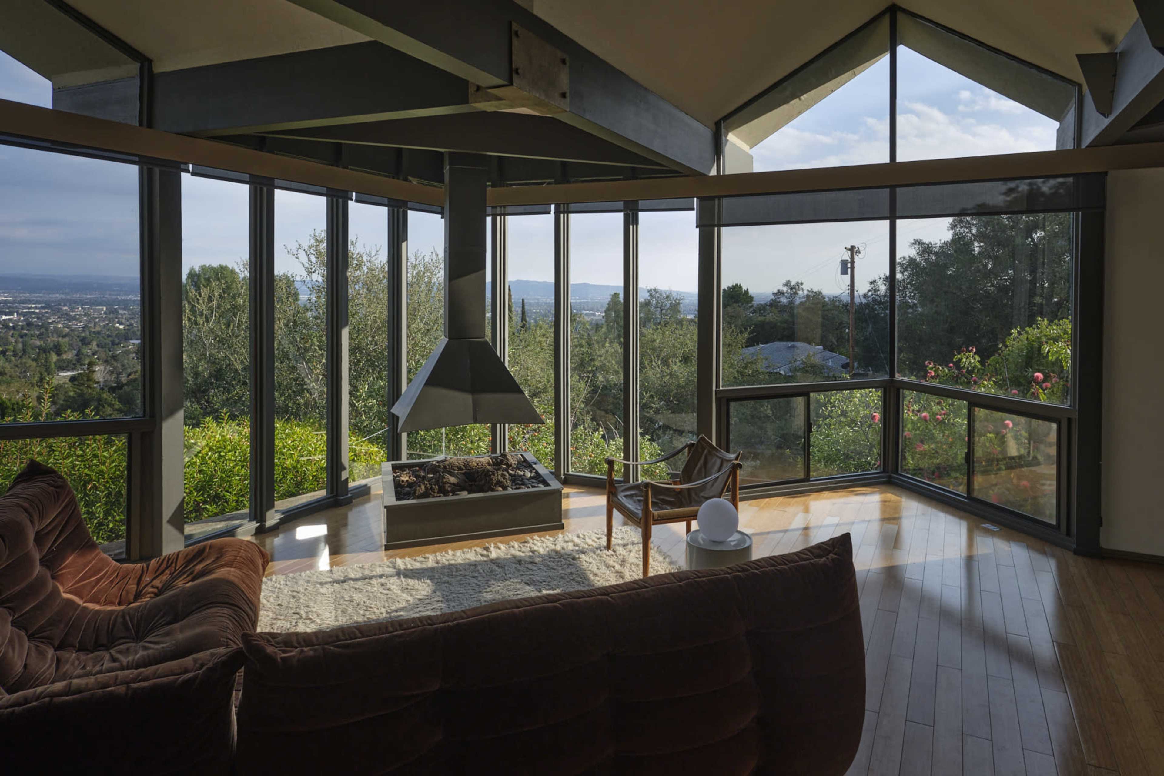 A modern living room with large windows offering a view of the outdoors, featuring a central fireplace and a brown sofa.