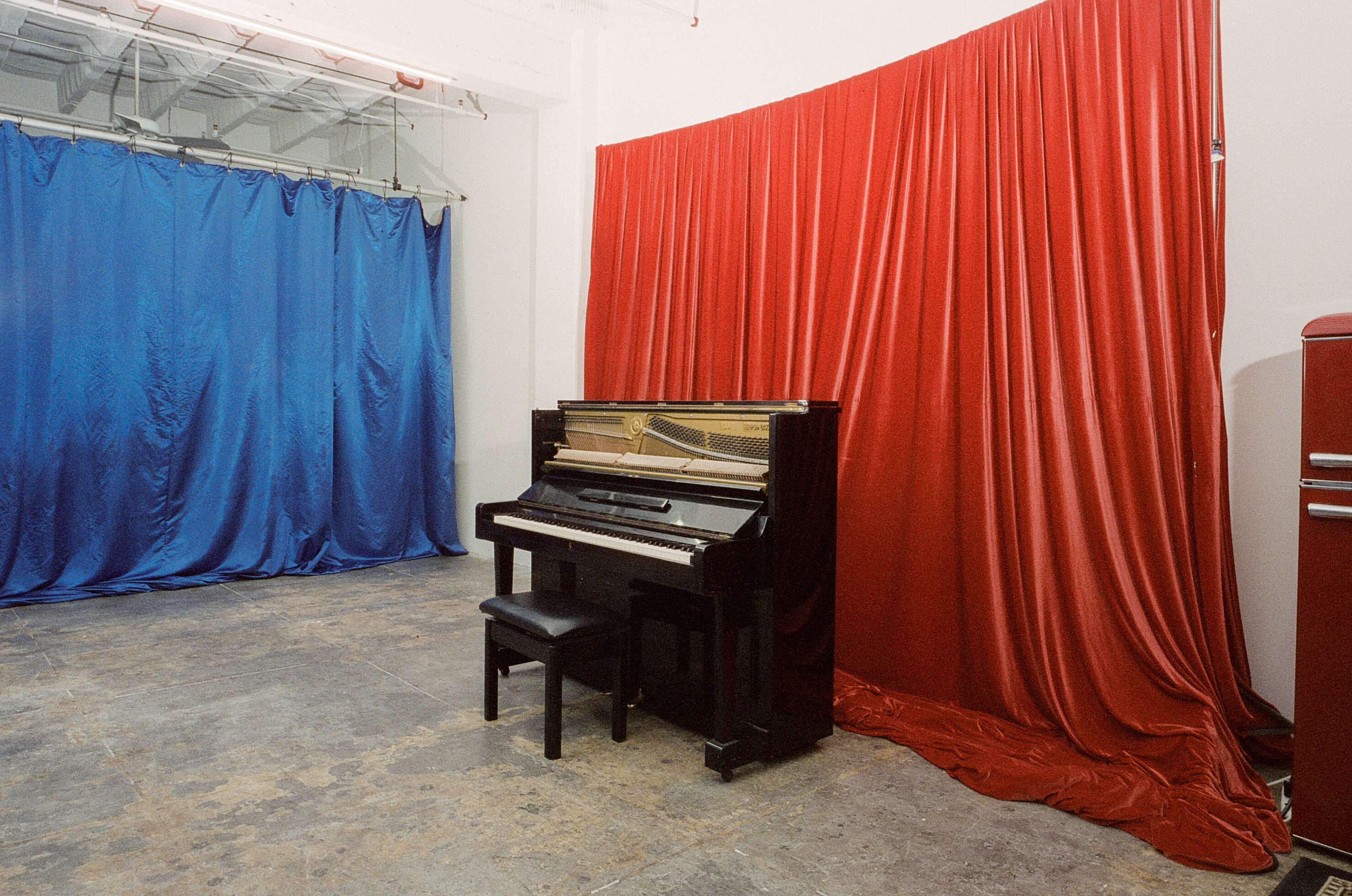 A black piano with a matching stool positioned against a red curtain, with blue drapes in the background.