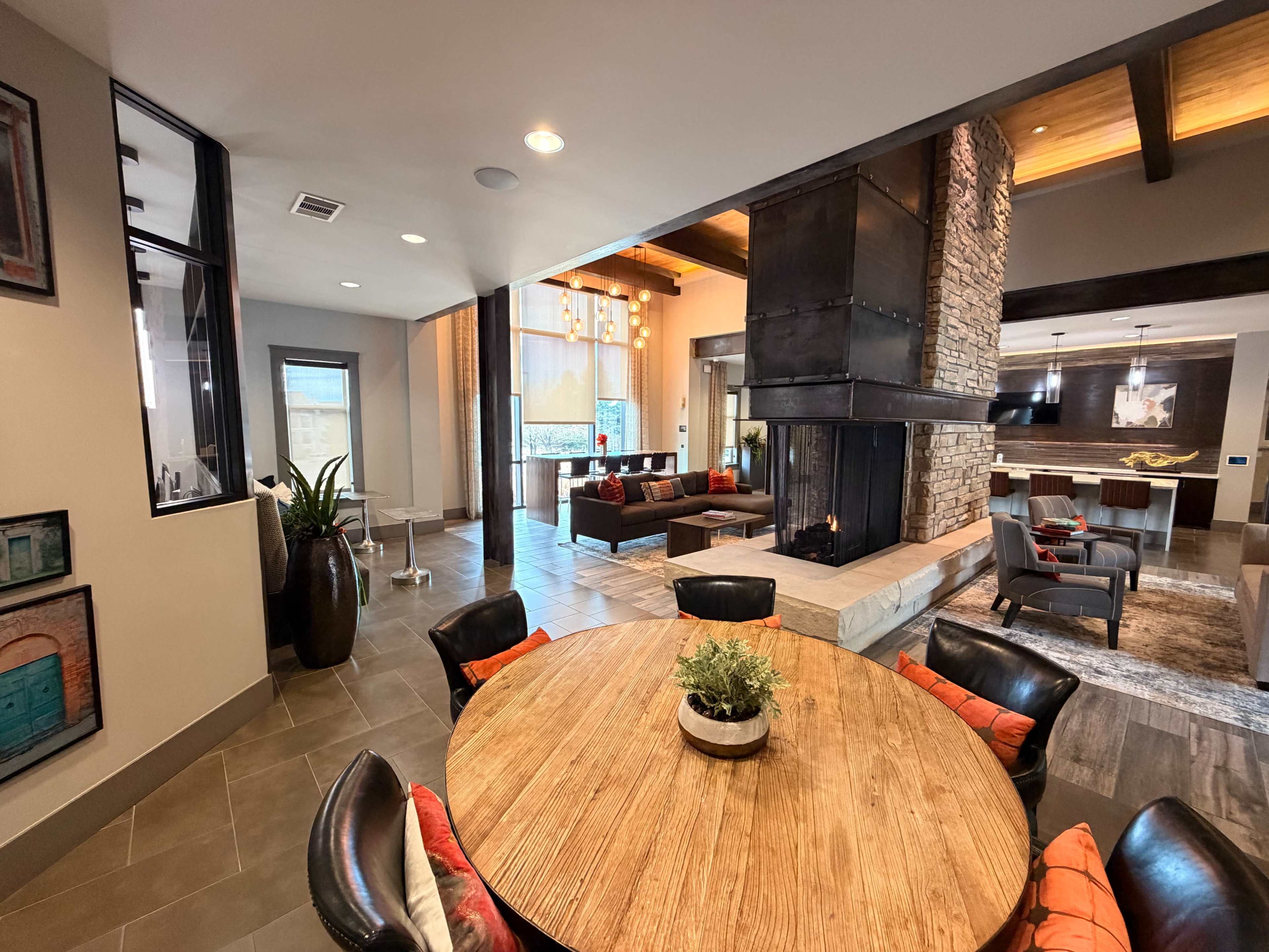 The image shows a modern living space featuring a round wooden dining table surrounded by black chairs, with a fireplace and a cozy seating area in the background.
