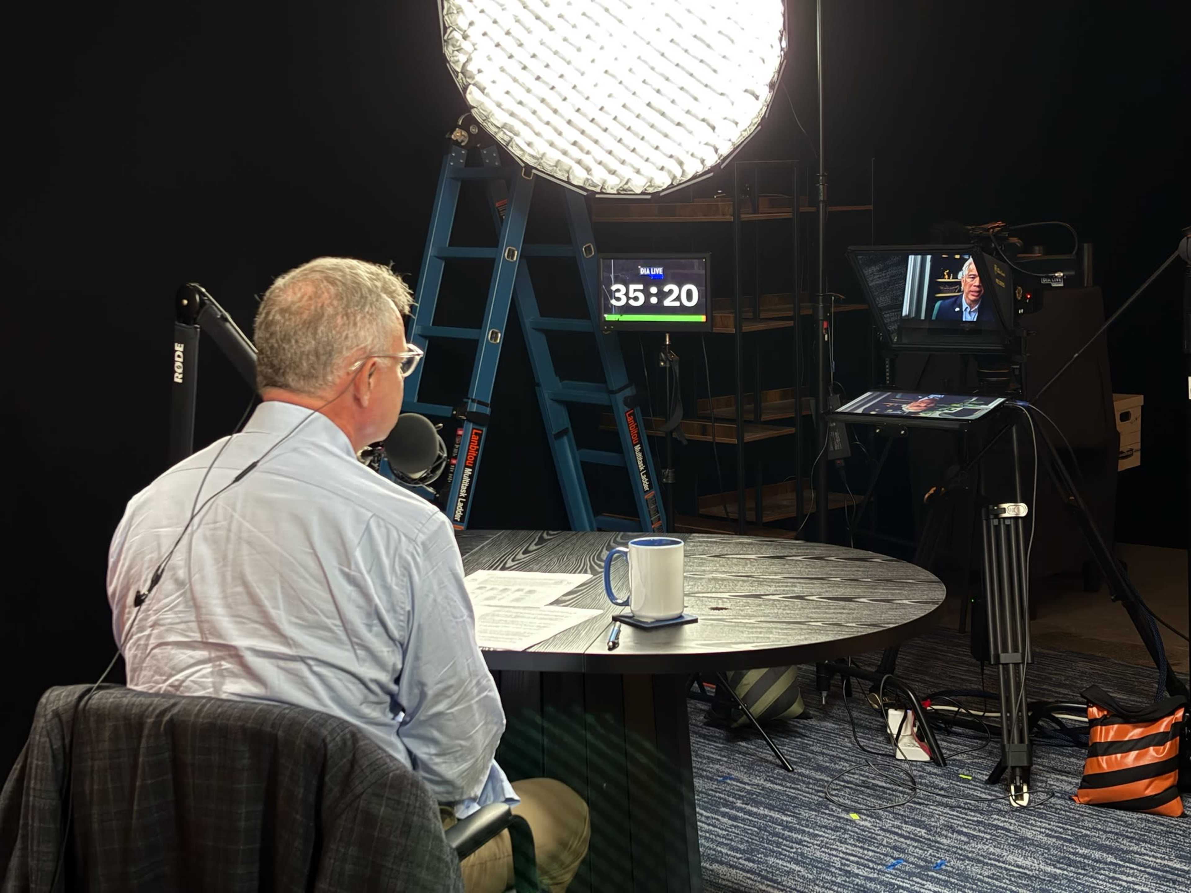 A man sits at a round table with a microphone and coffee mug, facing a camera and a digital timer in a dimly lit studio.