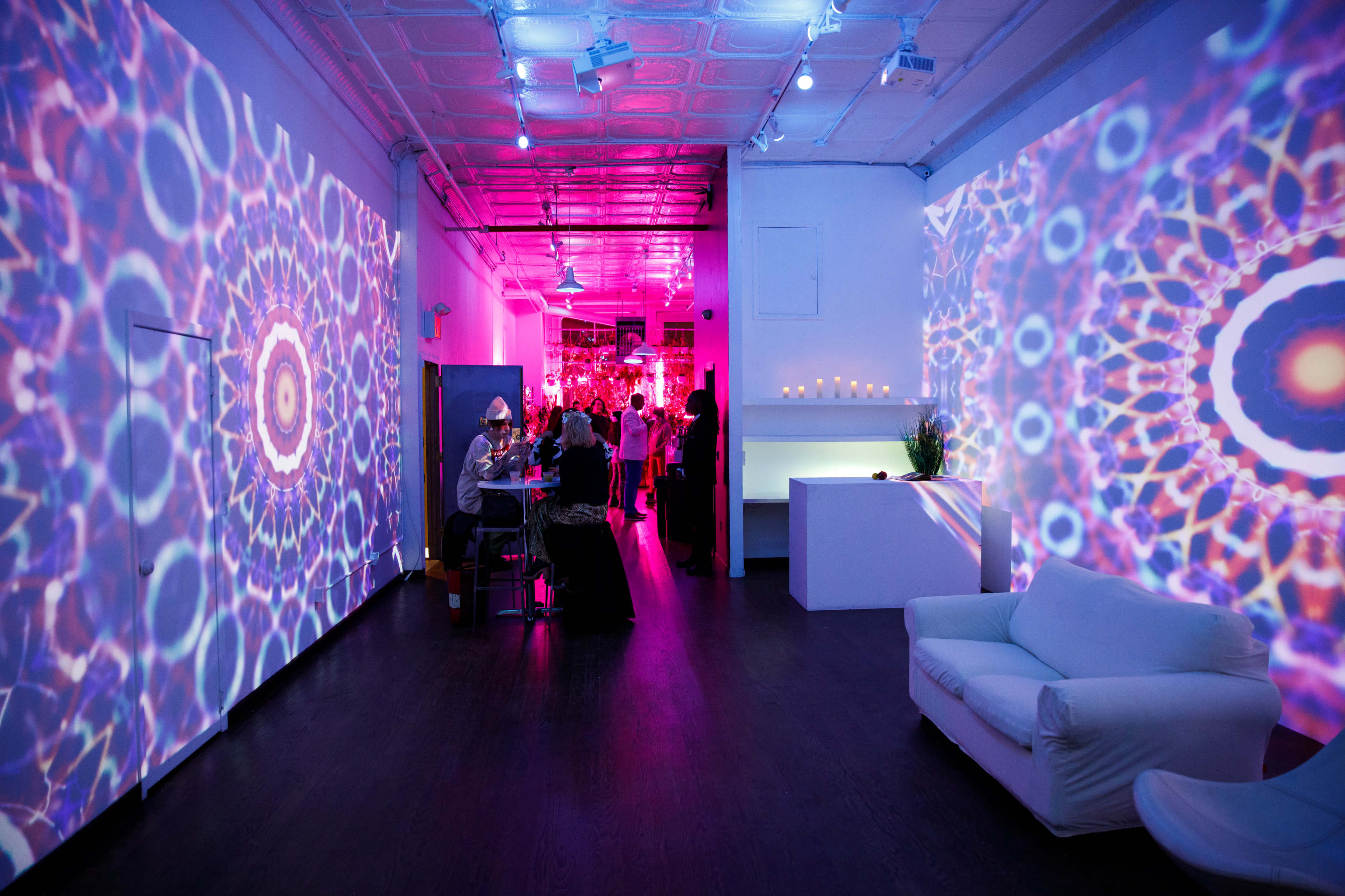 A brightly lit modern interior space with colorful, patterned projections on the walls, where guests are mingling near a white couch and a long table.