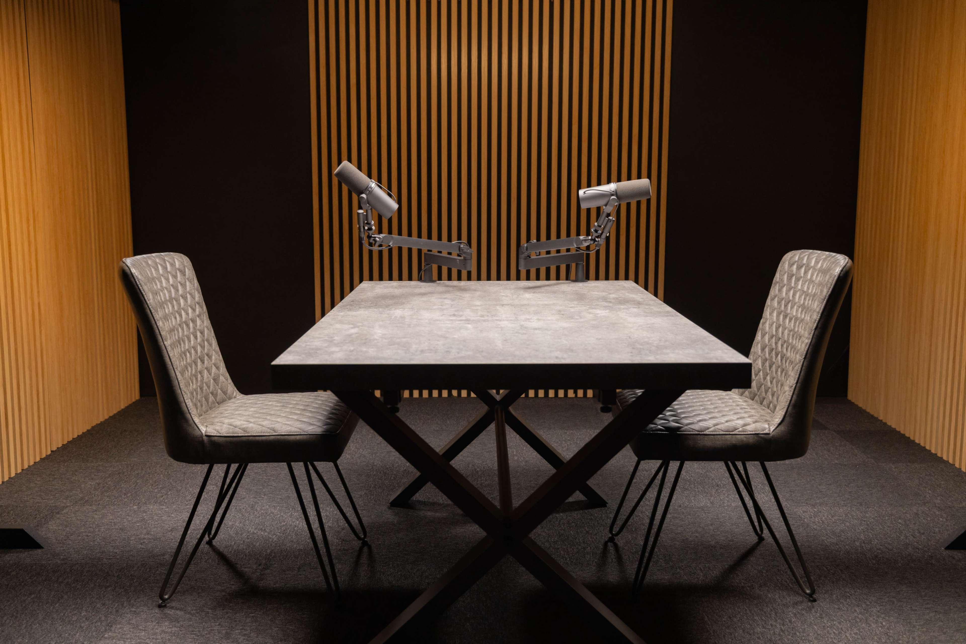 A modern recording studio features a concrete table with two chairs, equipped with microphone arms, set against a backdrop of wooden slats.