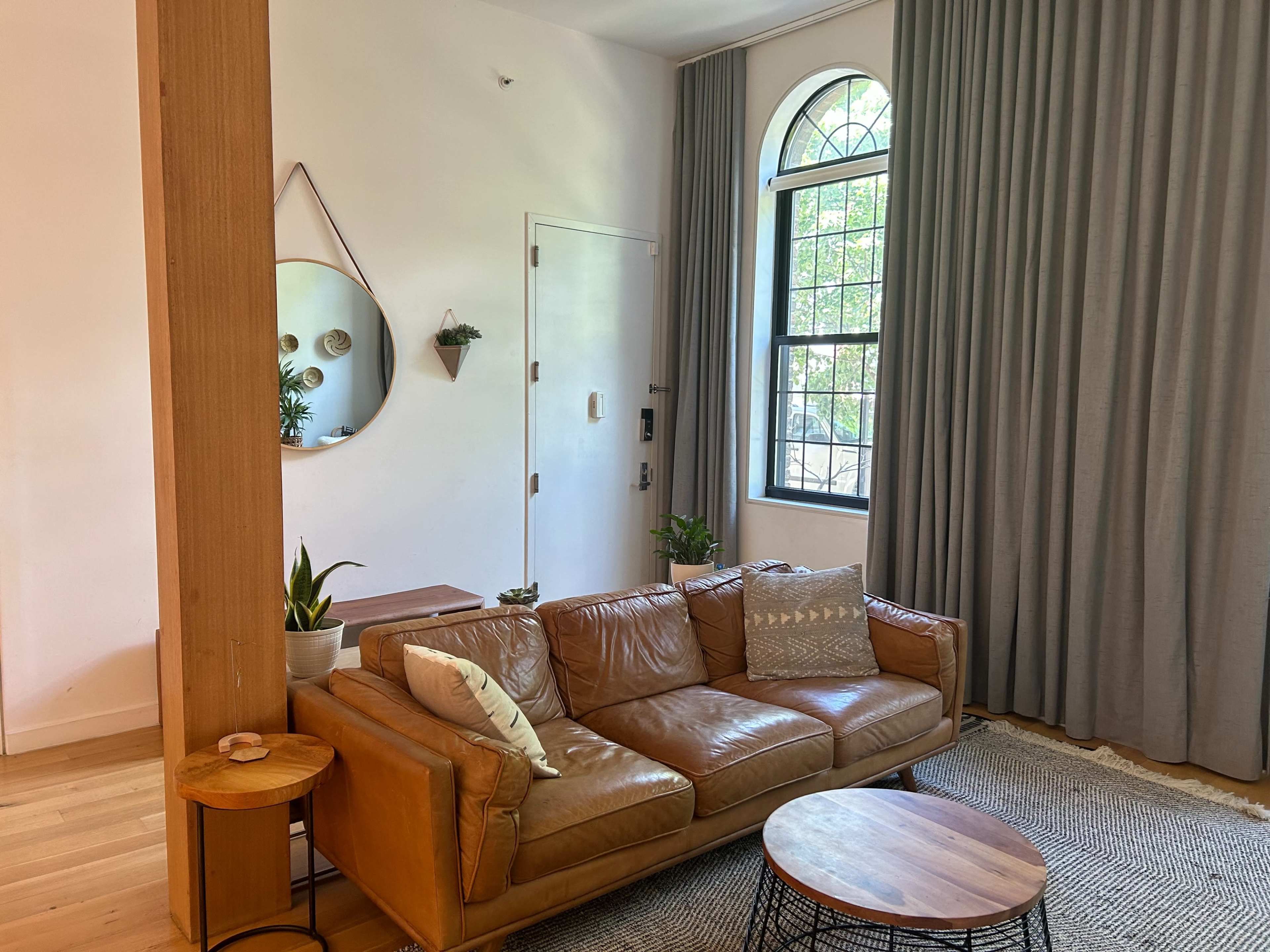 A cozy living room with a brown leather sofa, a round mirror on the wall, and large windows draped with gray curtains.