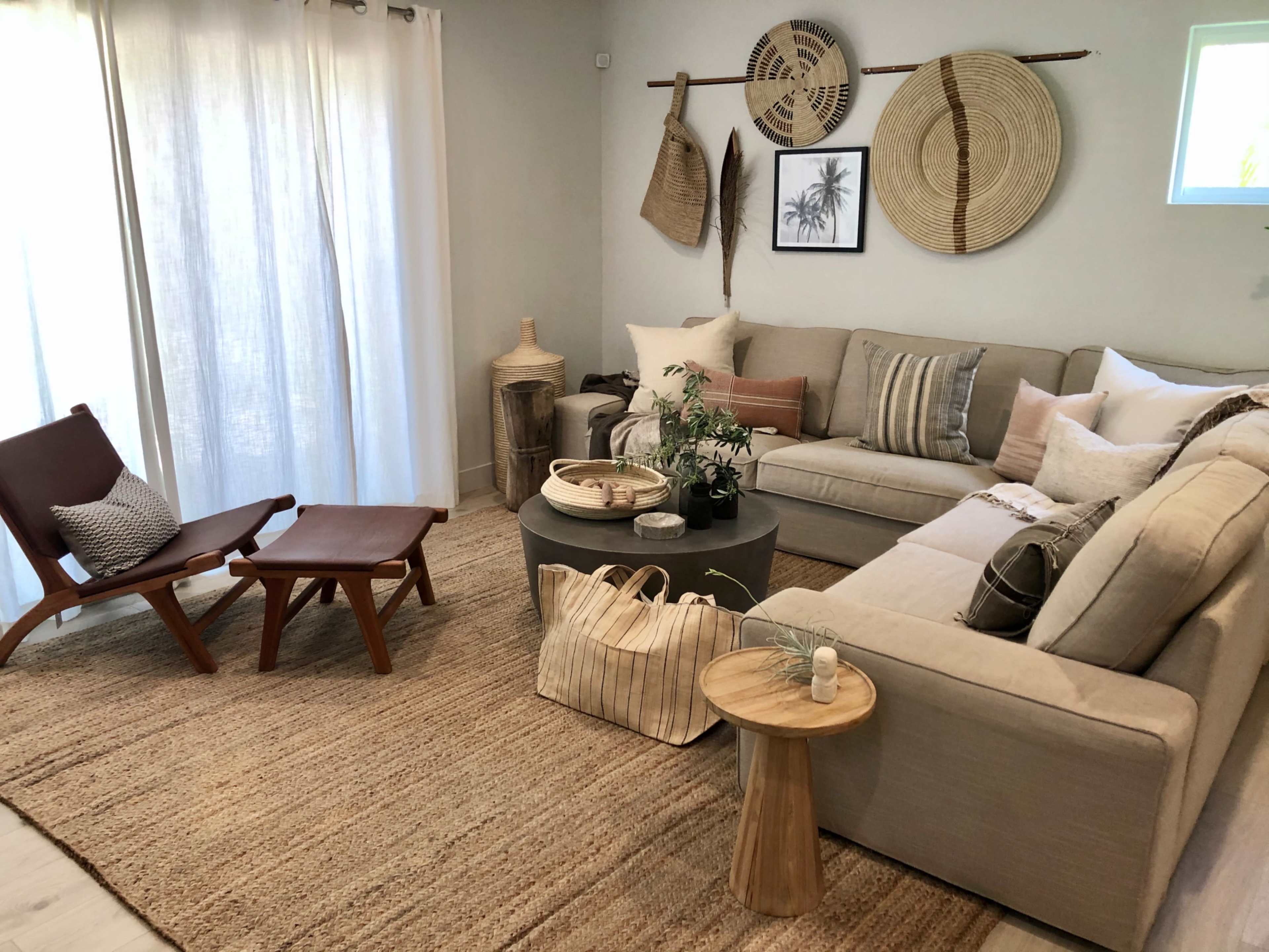 The image shows a light, modern living room with a sectional sofa, a coffee table, and a chair, surrounded by natural decor elements and a woven rug.