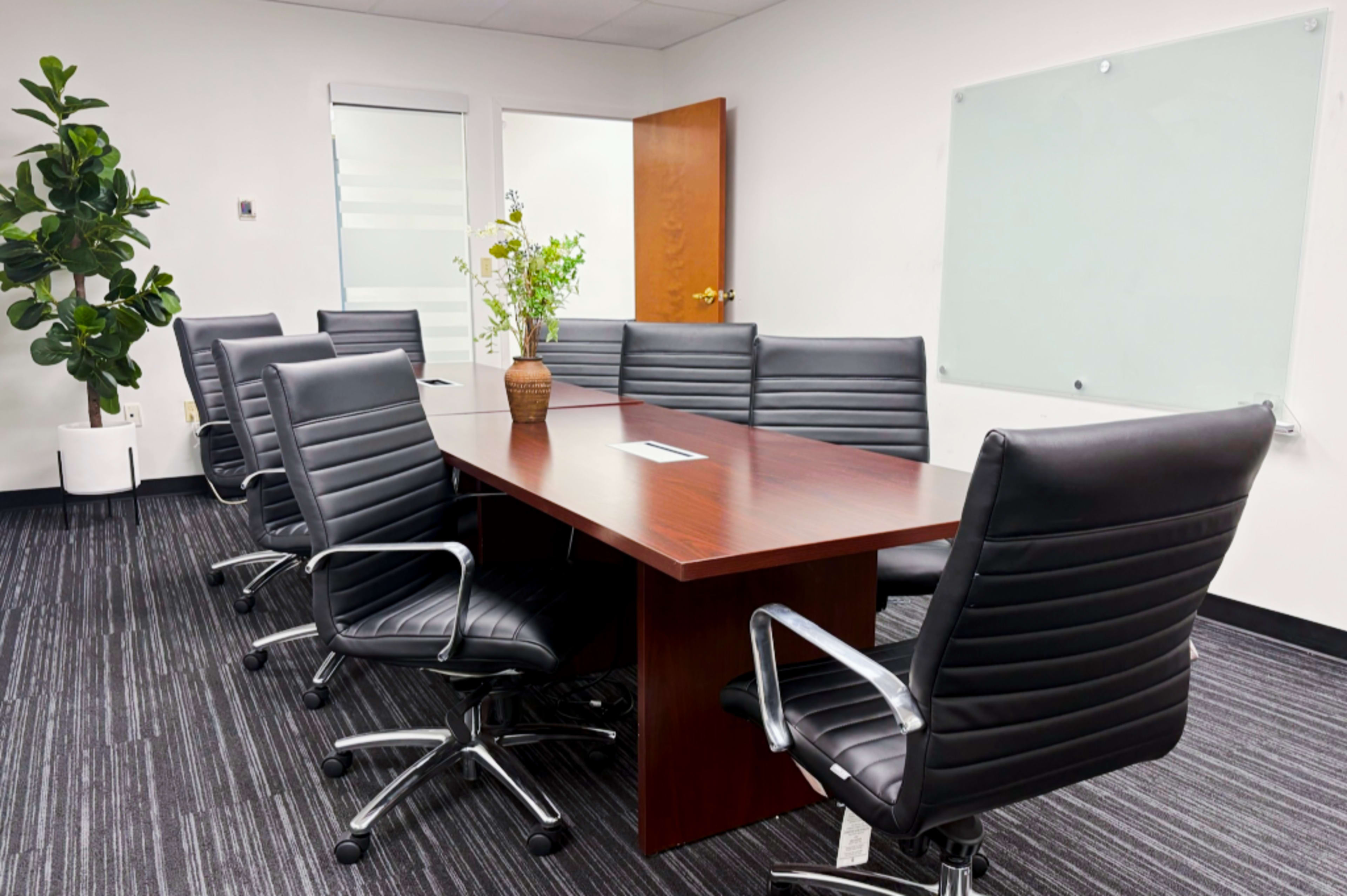 A modern conference room features a large wooden table surrounded by eight black leather chairs, with a potted plant and a whiteboard in the background.