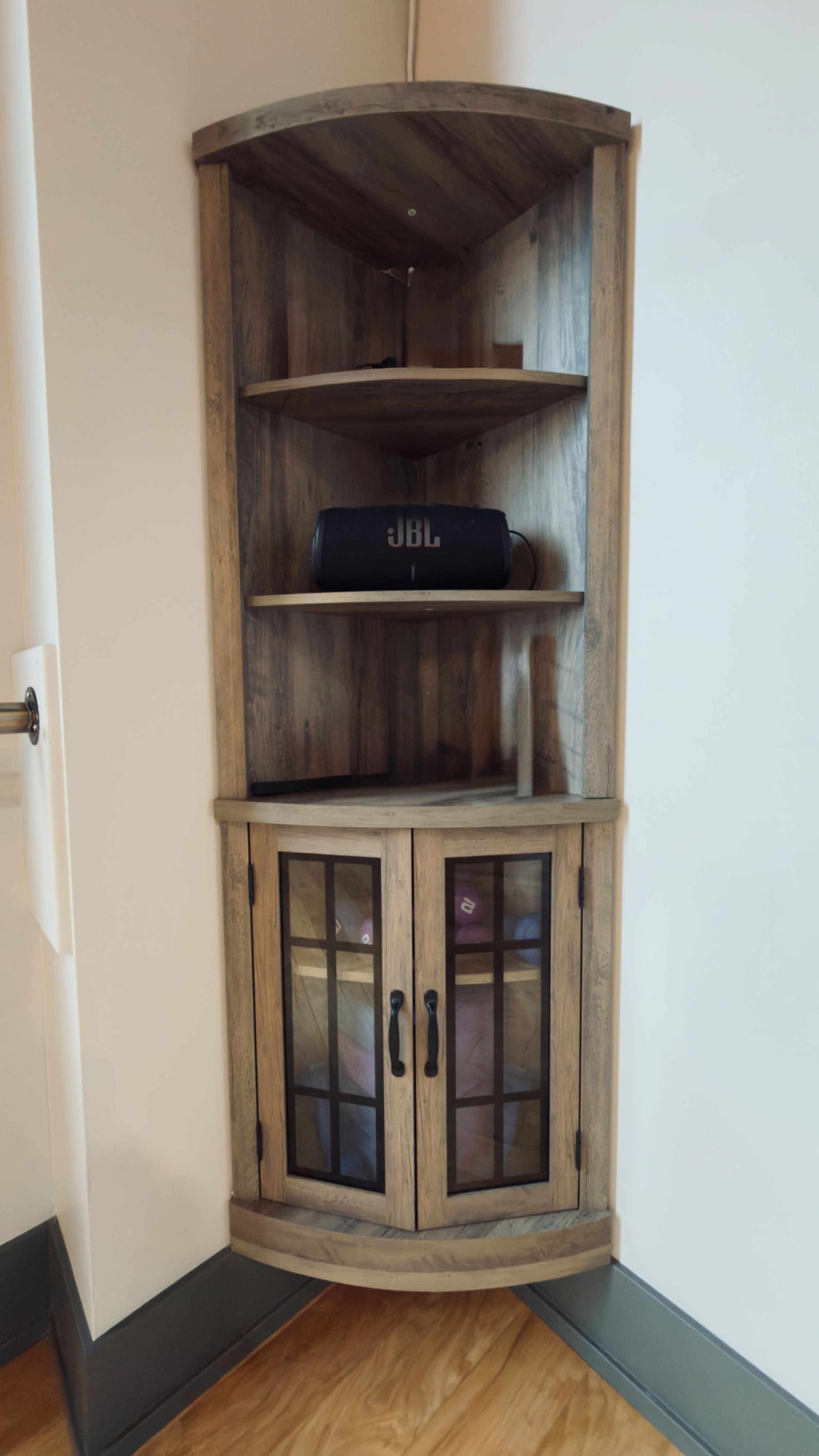 A wooden corner bookshelf with three open shelves and two glass-paneled doors at the bottom is positioned against a wall.