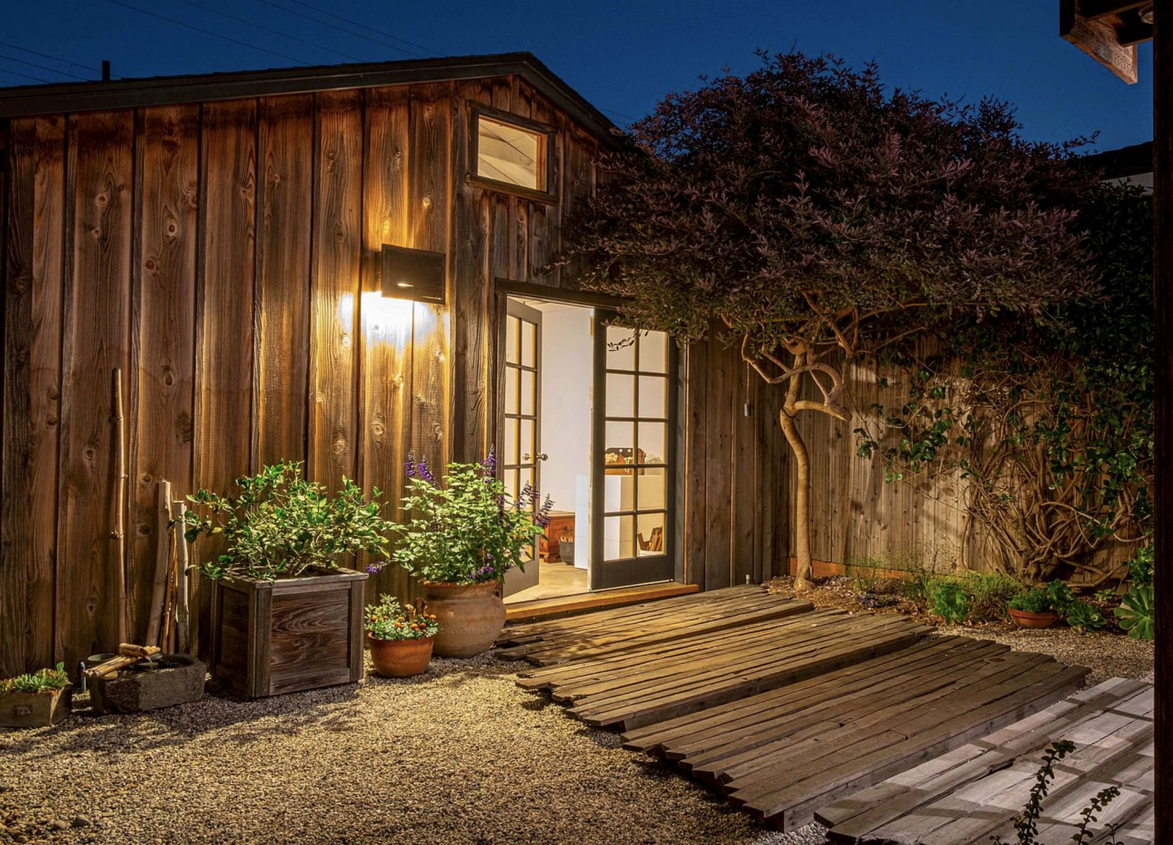 A wooden building with a lighted entrance and a garden area featuring plants and wooden walkways at dusk.