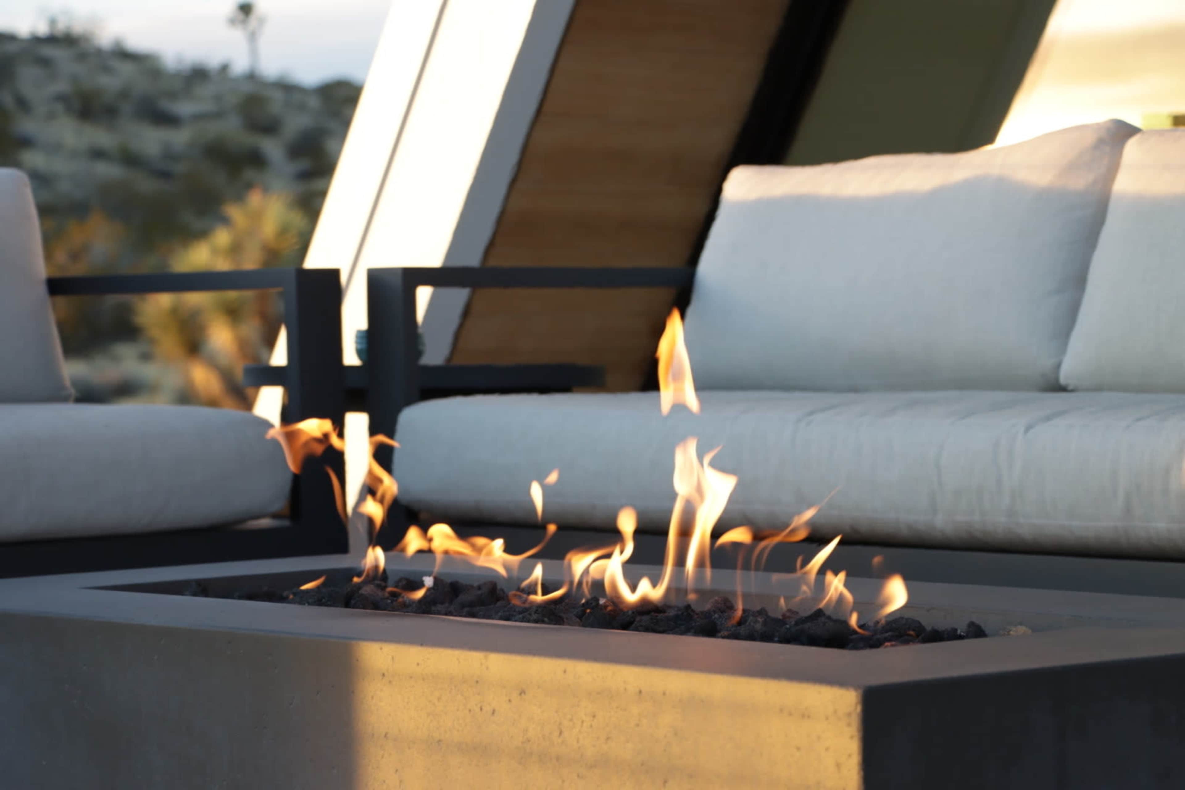 A modern outdoor seating area features a rectangular fire pit with flames and a stone base, surrounded by minimalist couches with light-colored cushions.