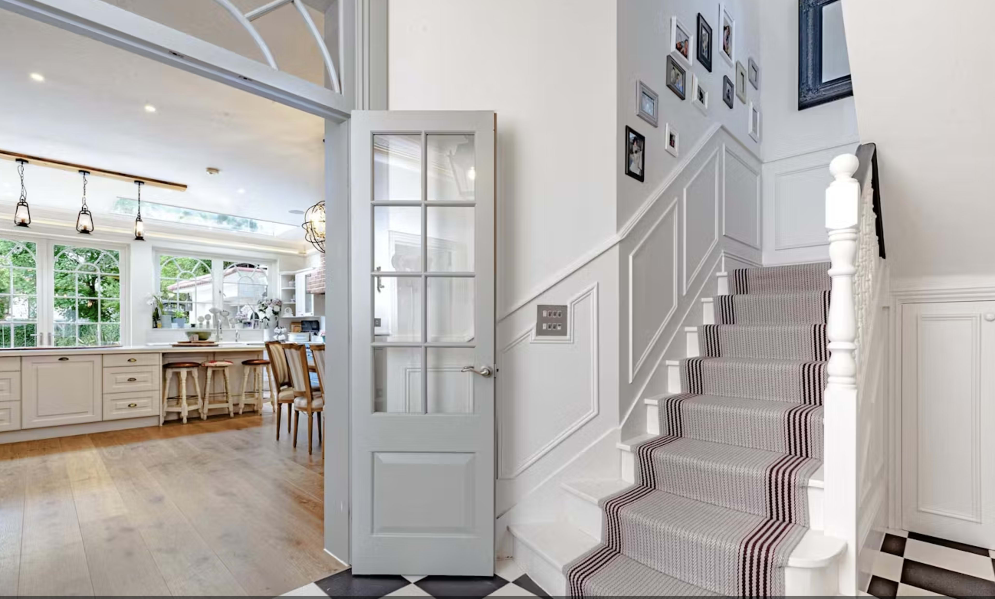 A light-filled entryway leads to a staircase with a patterned runner, adjacent to an open kitchen and dining area visible through a glass door.