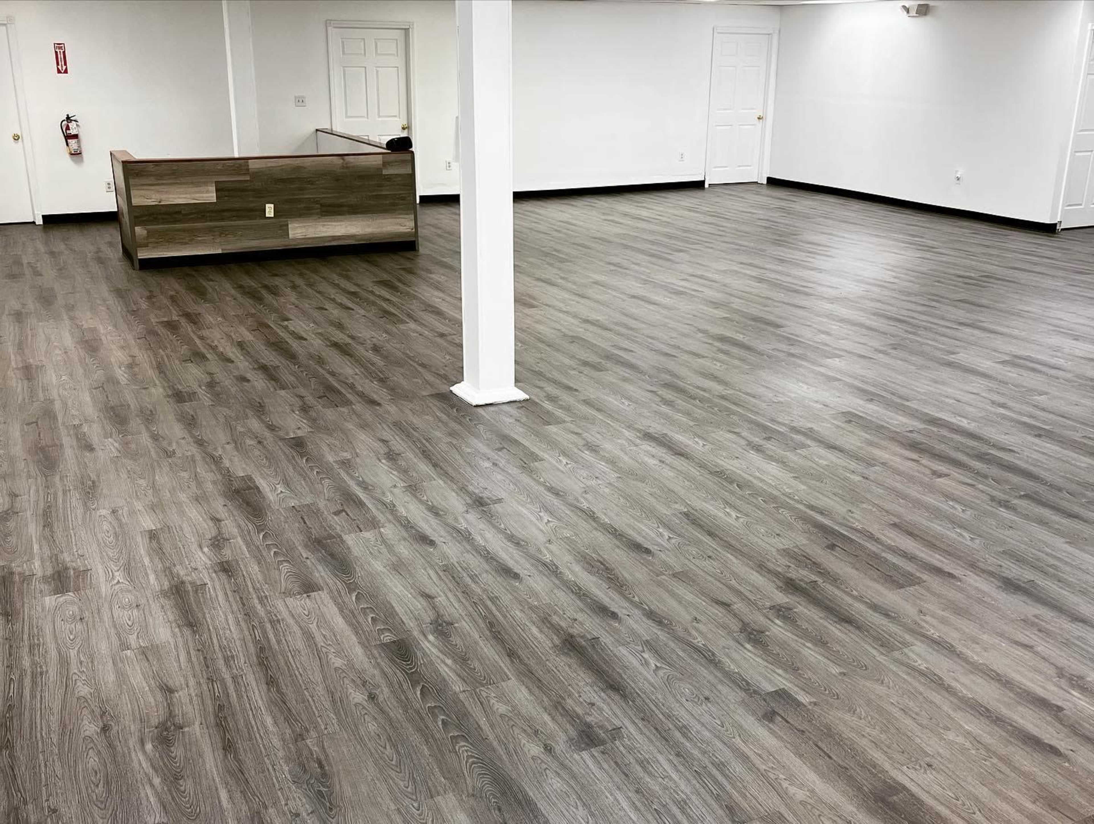 A spacious interior with wooden flooring and minimal furnishings, featuring a reception area in the background.