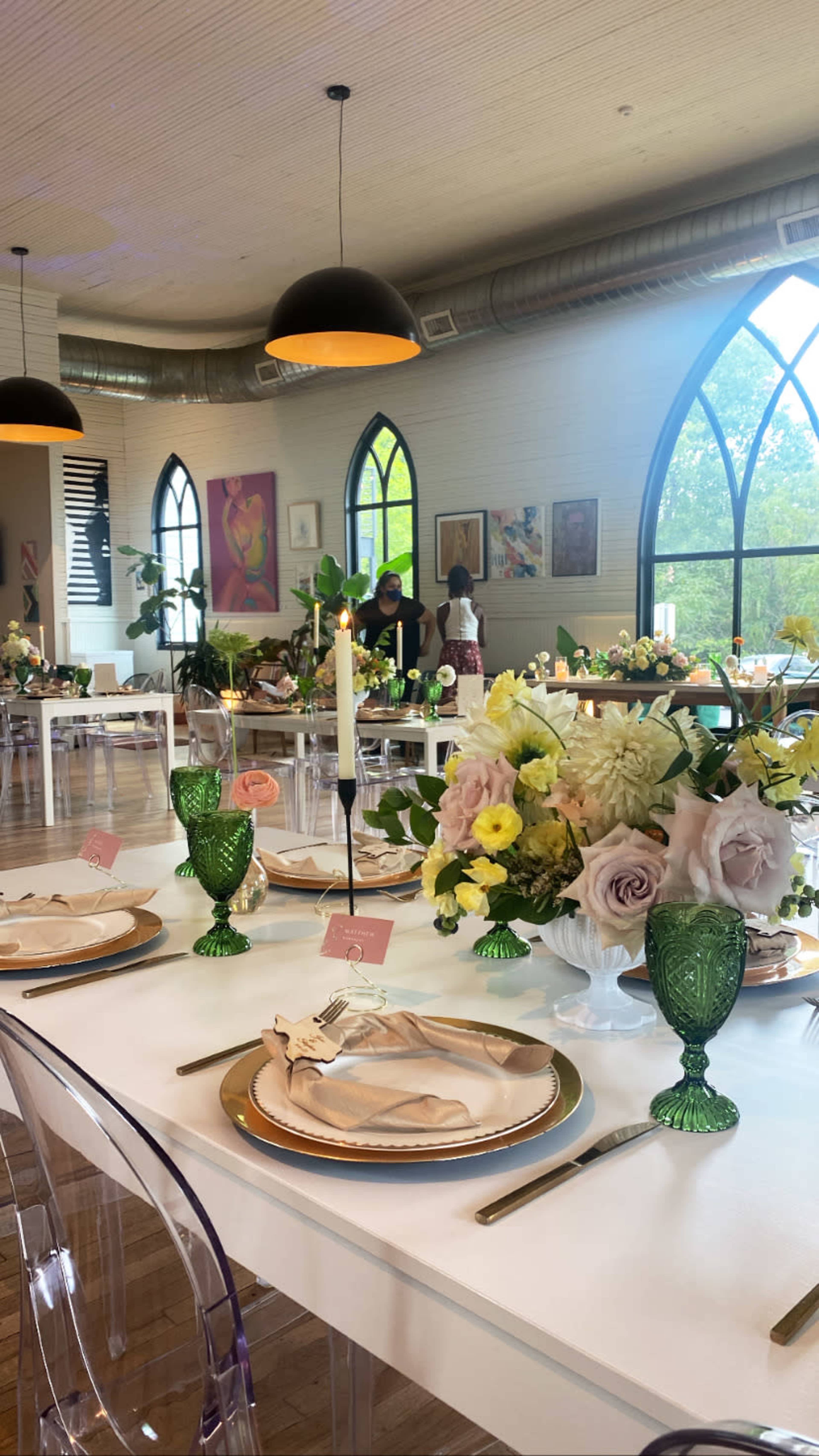 A dining area is set with elegantly arranged tables featuring floral centerpieces, decorative glassware, and place settings, with windows allowing natural light to illuminate the space.