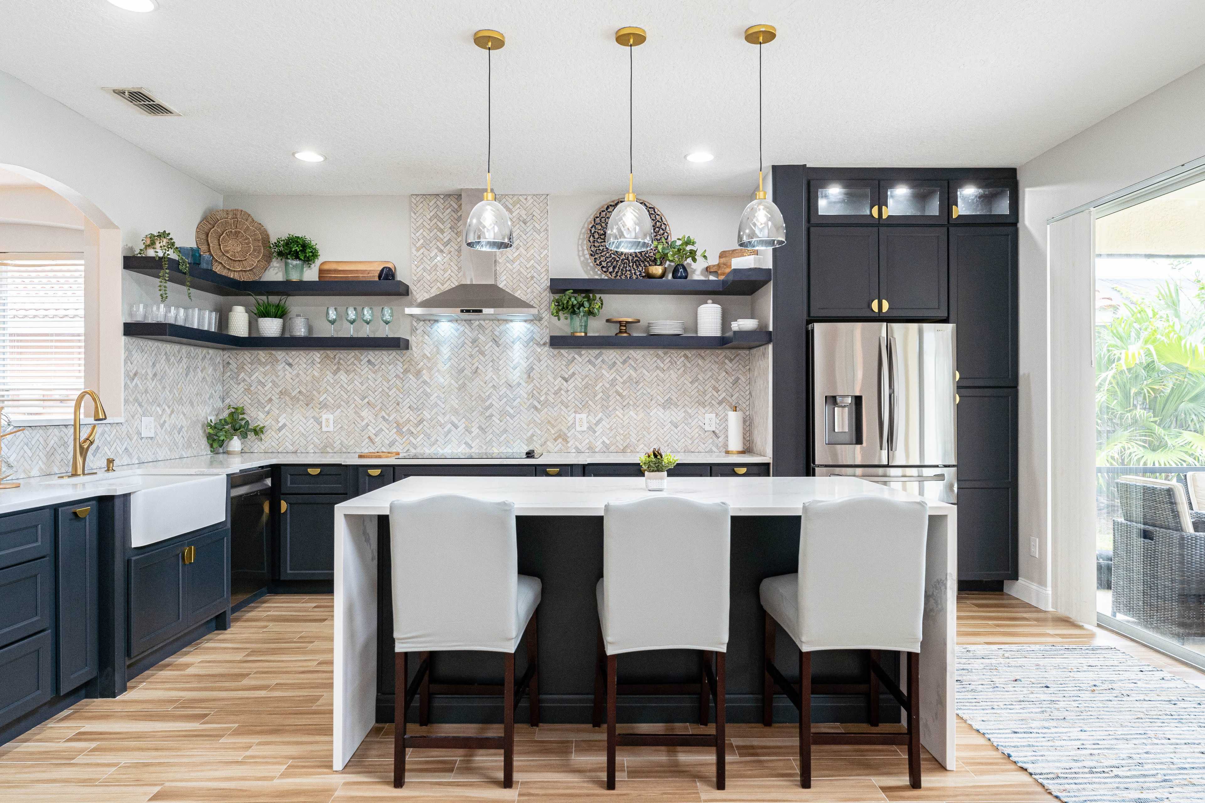 The image shows a modern kitchen with dark cabinetry, a white countertop, and three upholstered bar stools at an island, featuring a patterned backsplash and various plants for decoration.