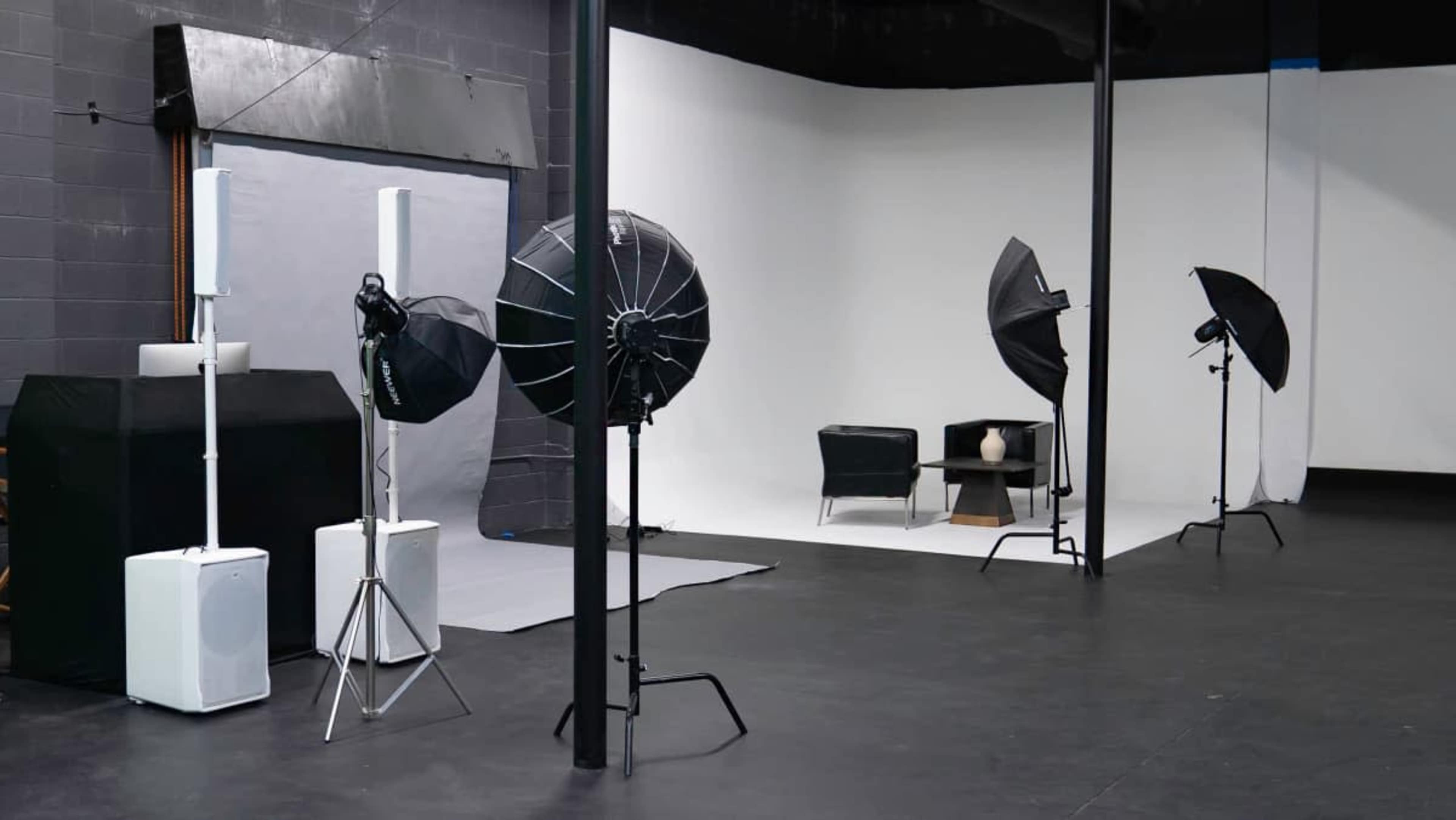 The image shows a photography studio with lighting equipment, a black chair, and a white backdrop.