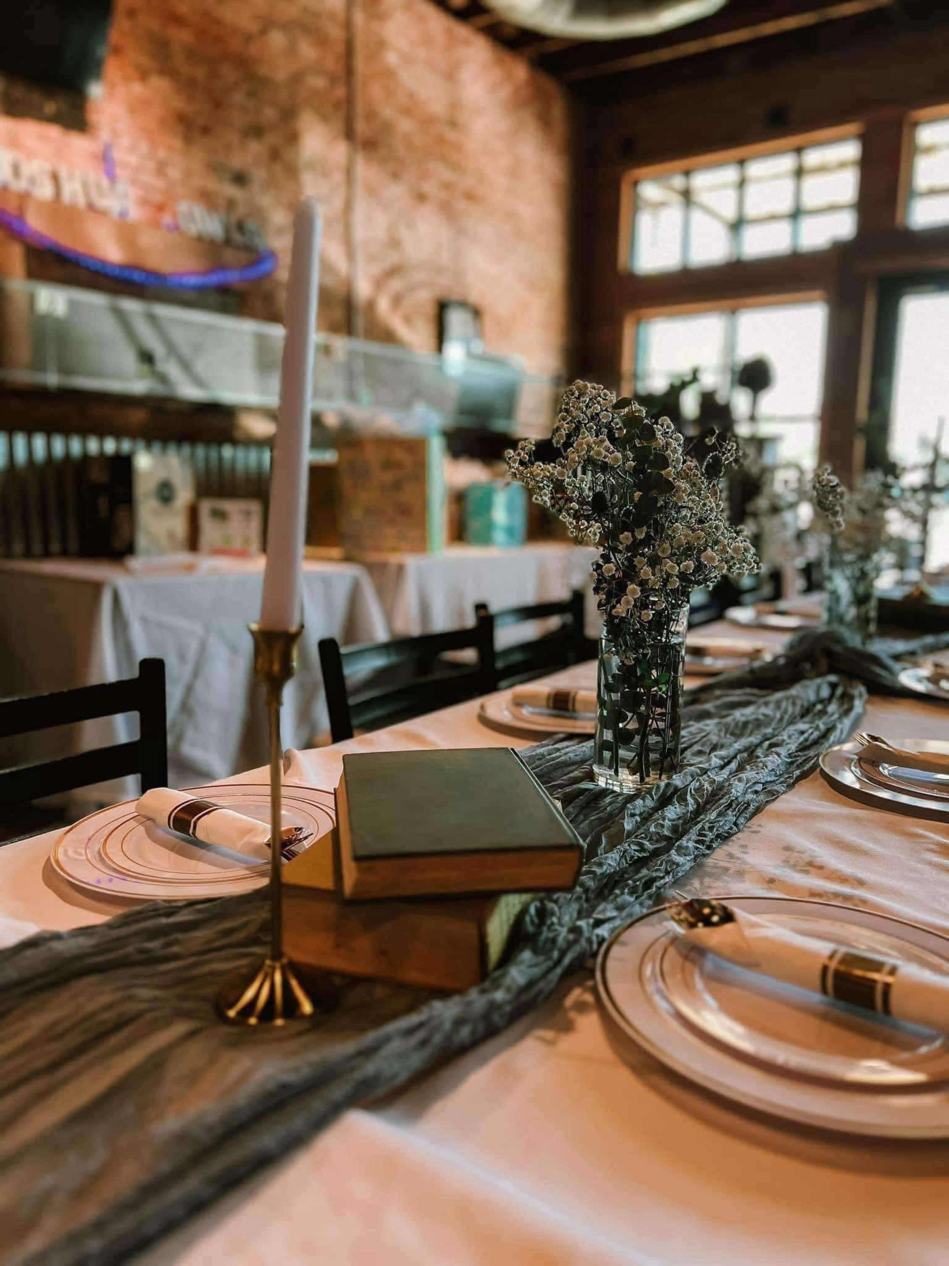 A table is set for an event with white plates, a gray table runner, a candle in a brass holder, a vase of small flowers, and stacked books as centerpieces.