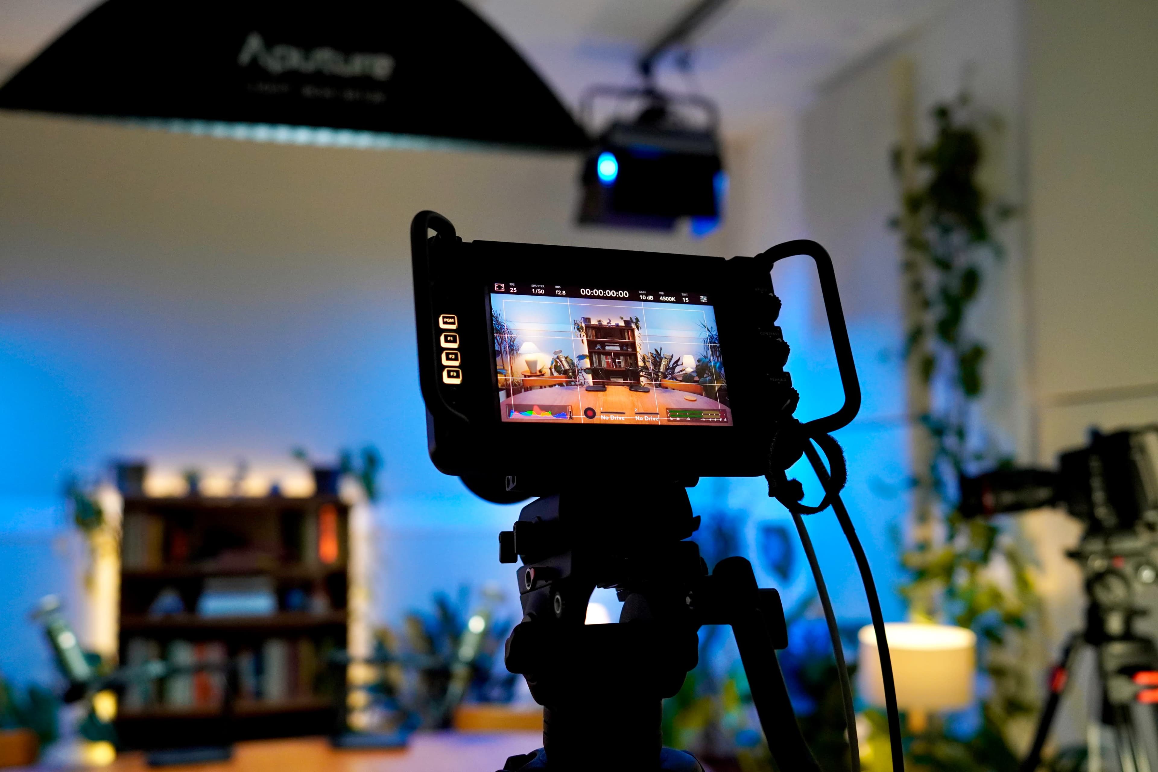 A camera on a tripod captures a well-lit indoor scene featuring a bookshelf and decorative plants.