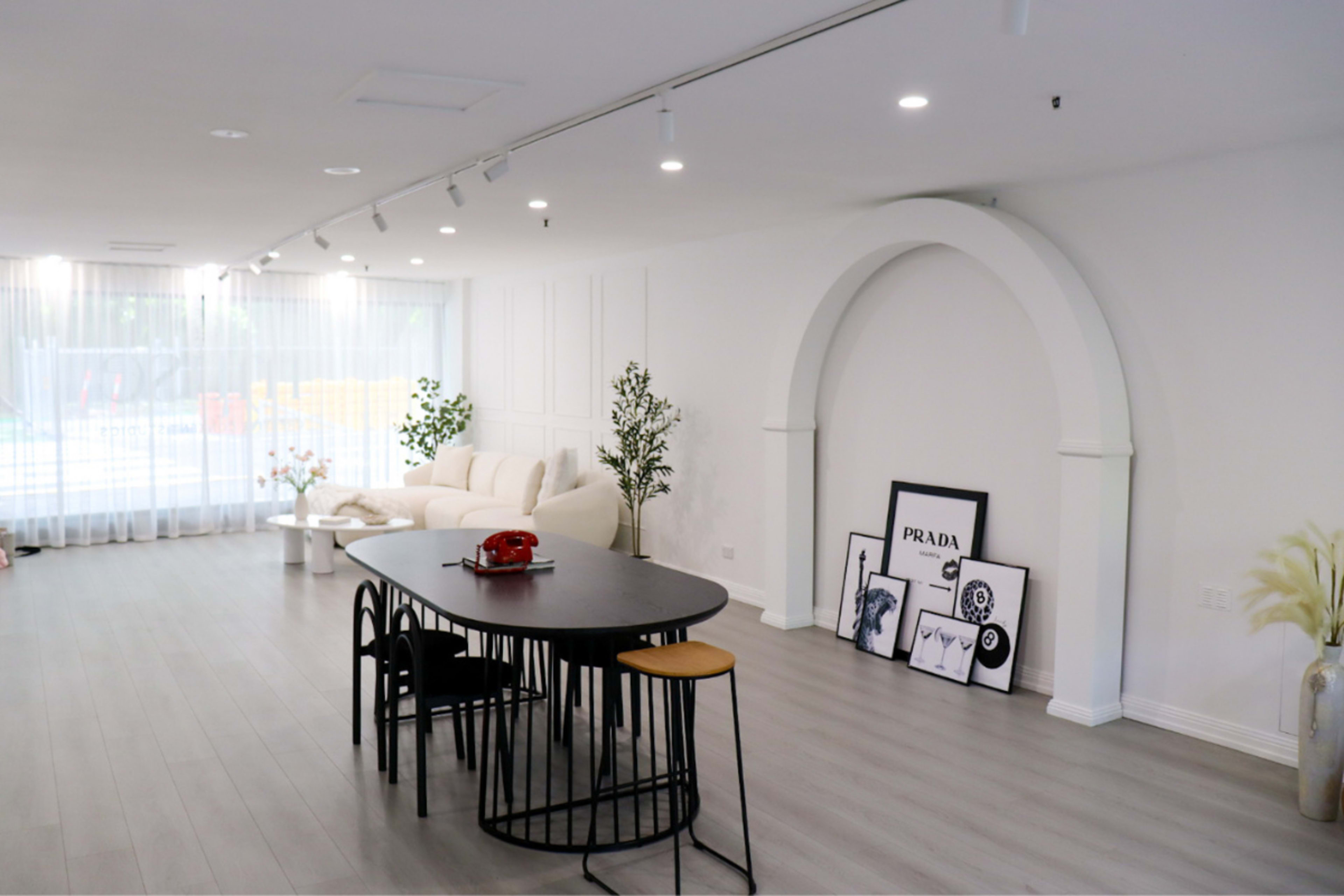 The scene features a modern interior space with a dining table surrounded by black chairs, a white sofa, and decorative plants, complemented by framed art on the floor.