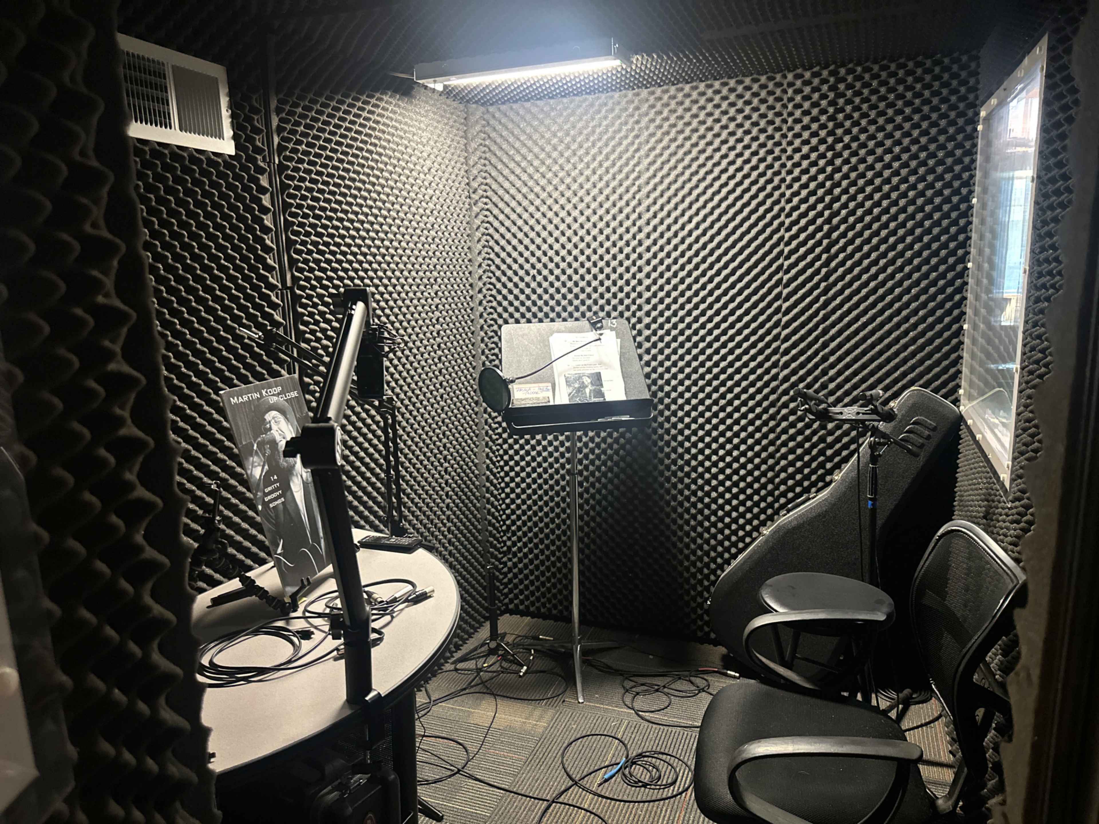 The image shows a soundproof recording studio with a desk, two chairs, a music stand, and acoustic foam lining the walls.