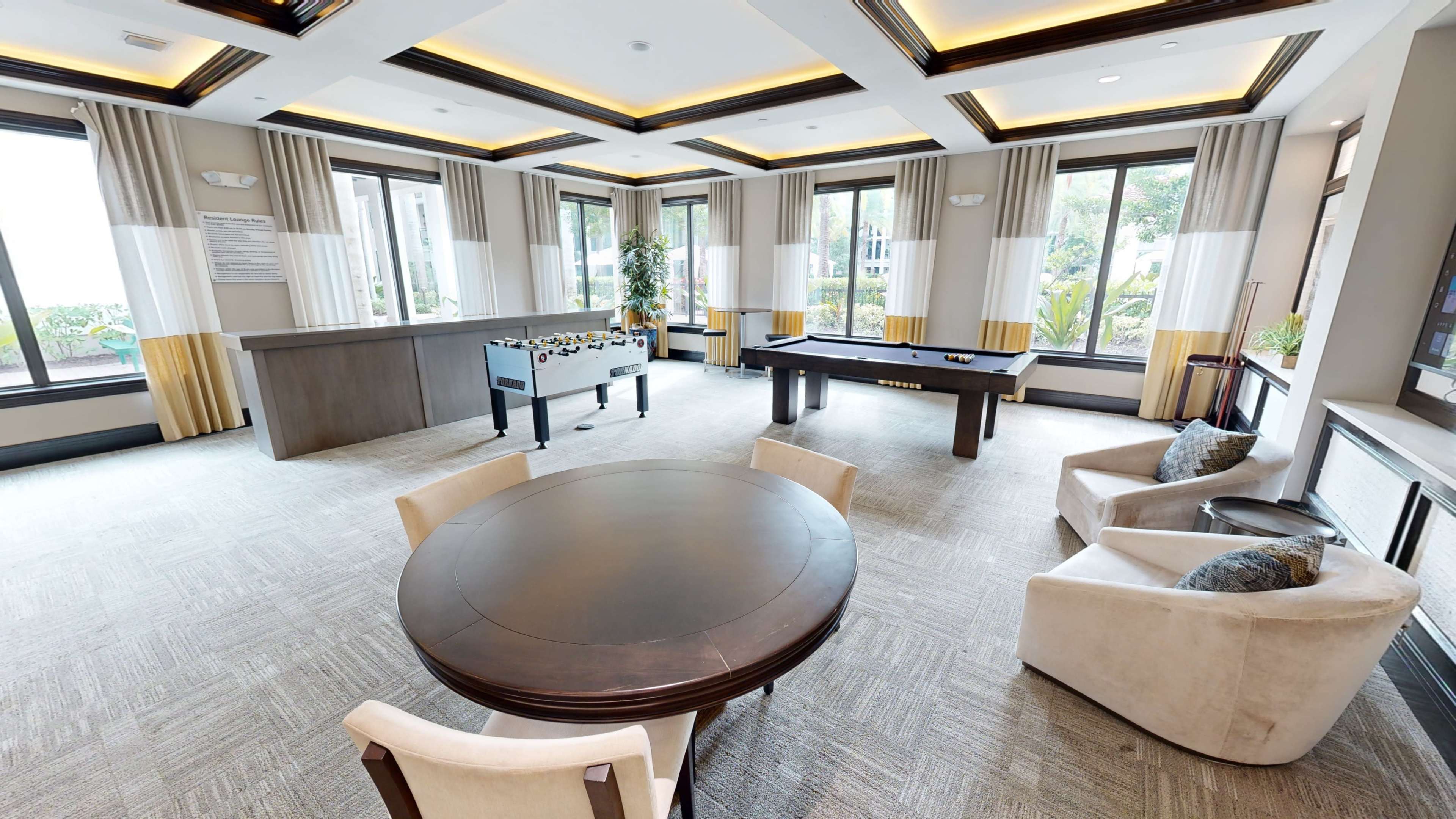 The image shows a spacious lounge area featuring a foosball table, a billiards table, a round dining table, and comfortable armchairs, with large windows letting in natural light.