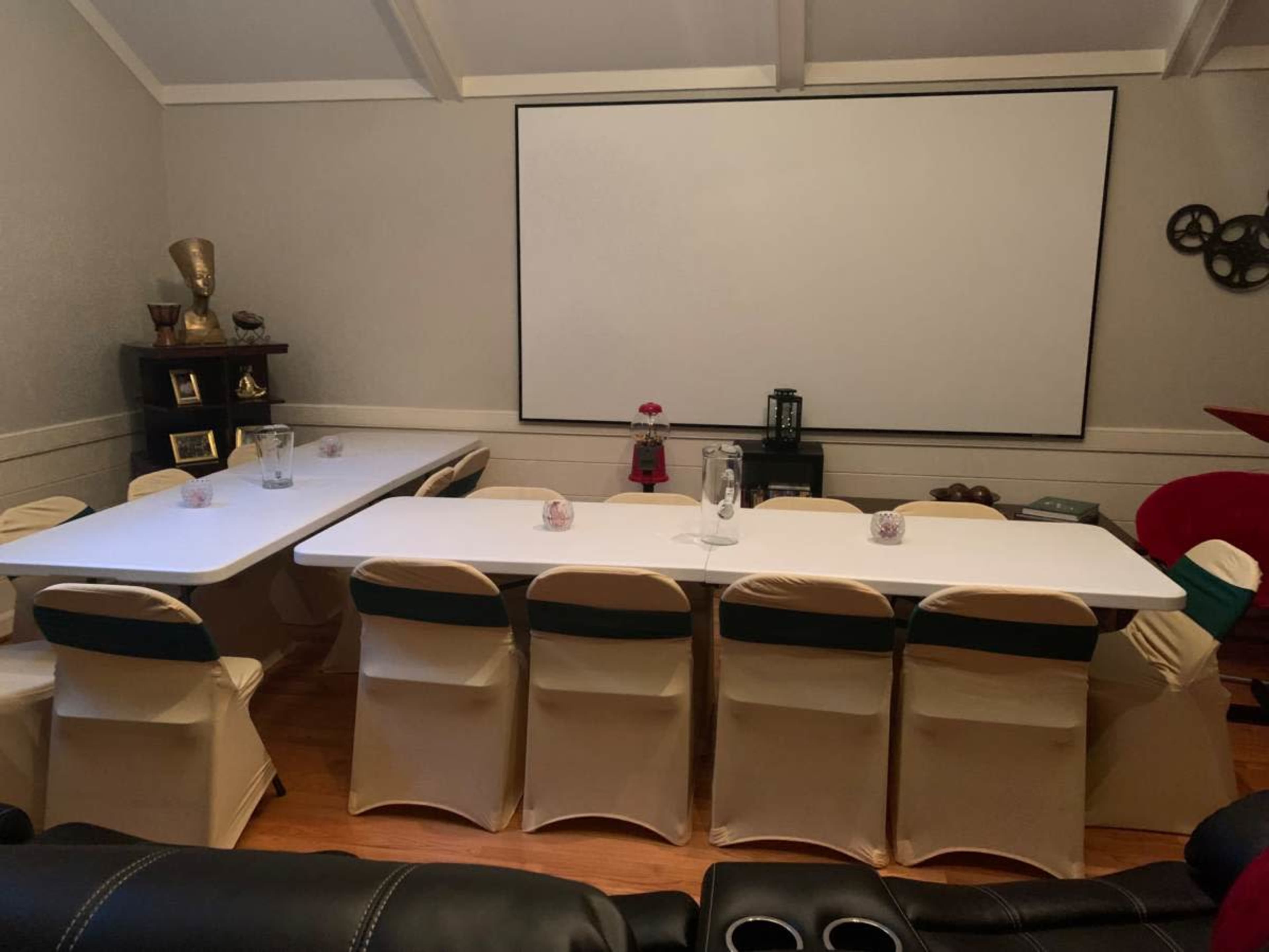 A conference room is set up with several long tables, chairs, and a large blank screen on the wall.