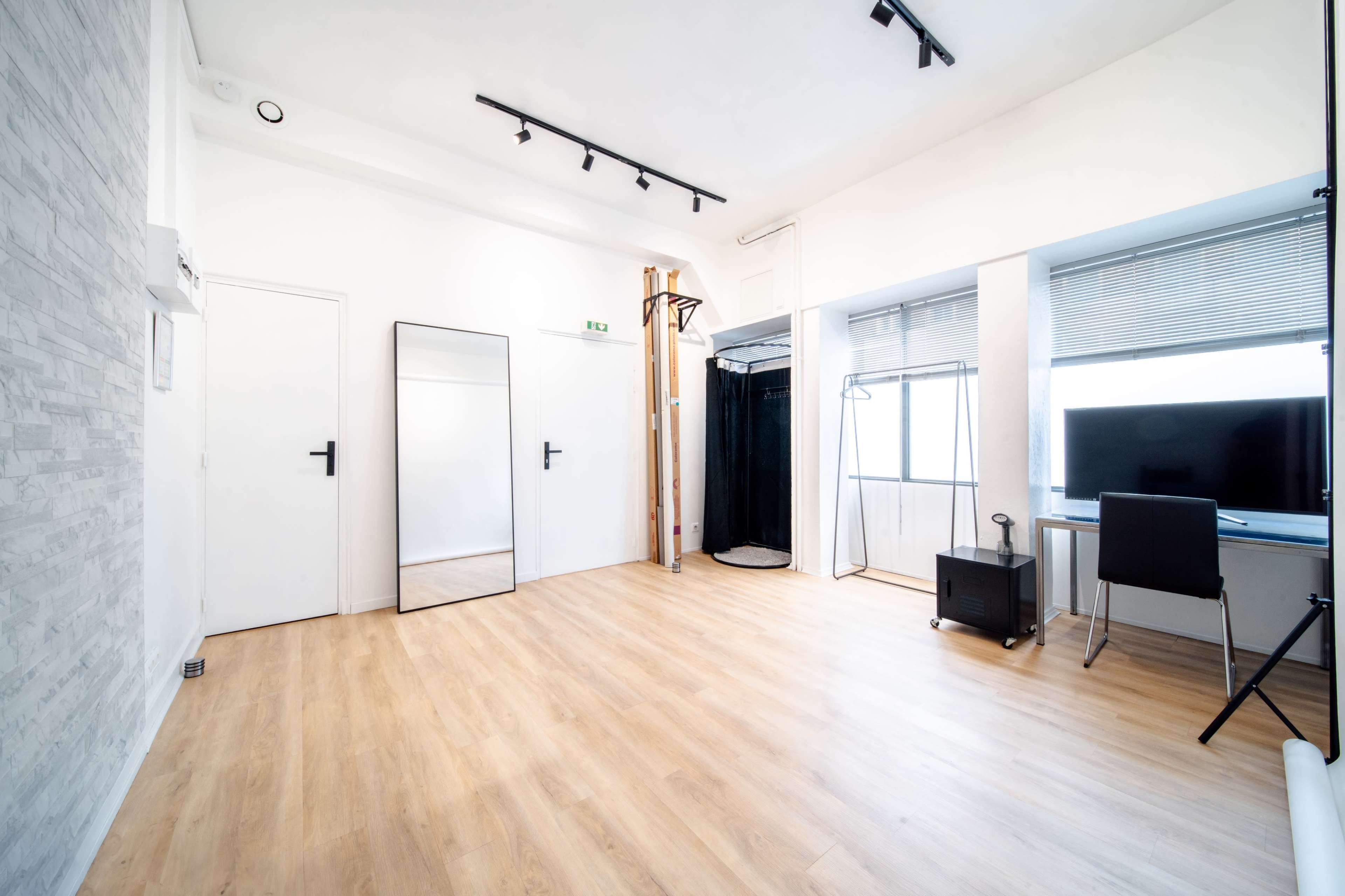 The image shows a bright, empty room with light wooden flooring, a large mirror, a TV, and a black clothing rack.