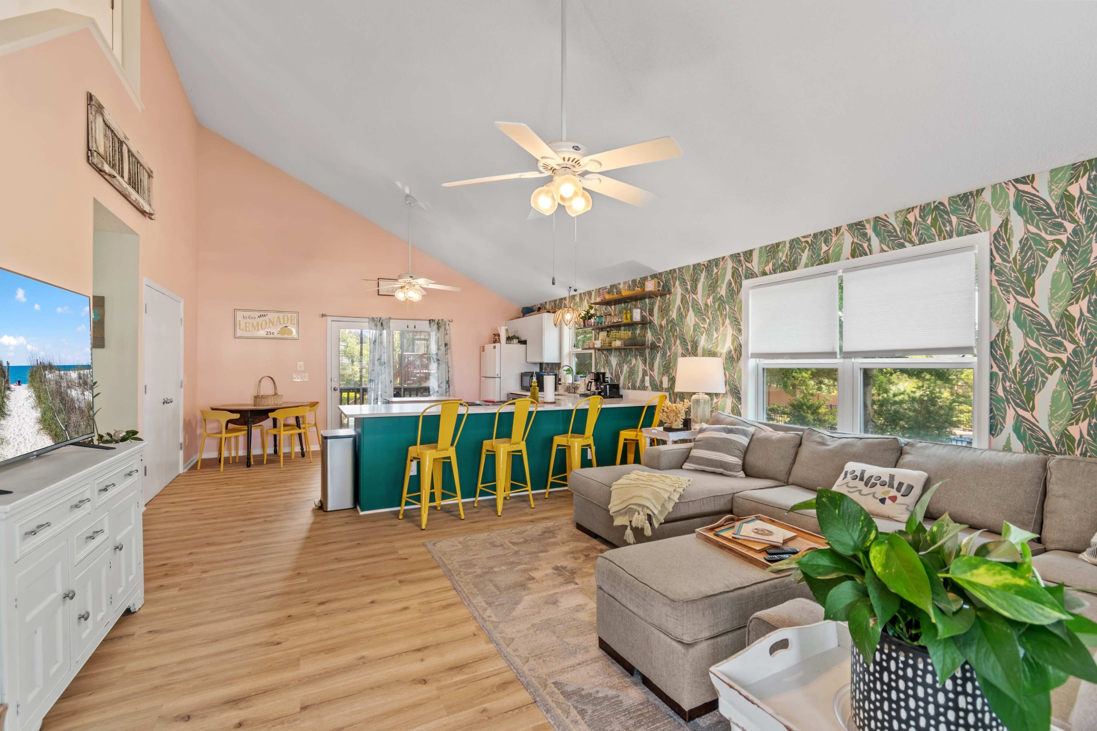 The image shows a bright, open living space with a sectional sofa, an island bar with yellow stools, and a kitchen in the background adorned with tropical-themed wallpaper.