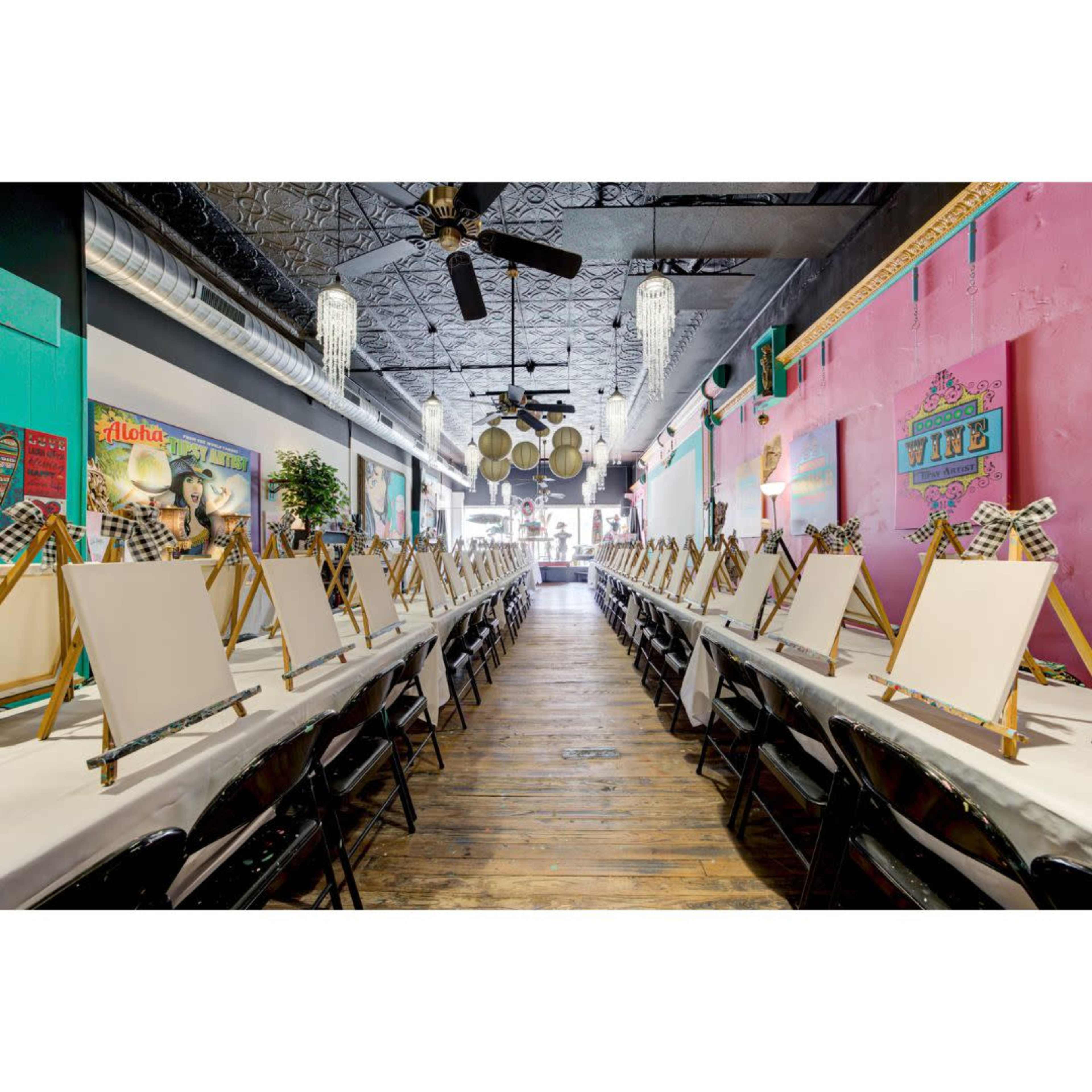 The image shows a spacious art studio with a long row of empty easels set up on tables, surrounded by colorful wall art and decorative lights.