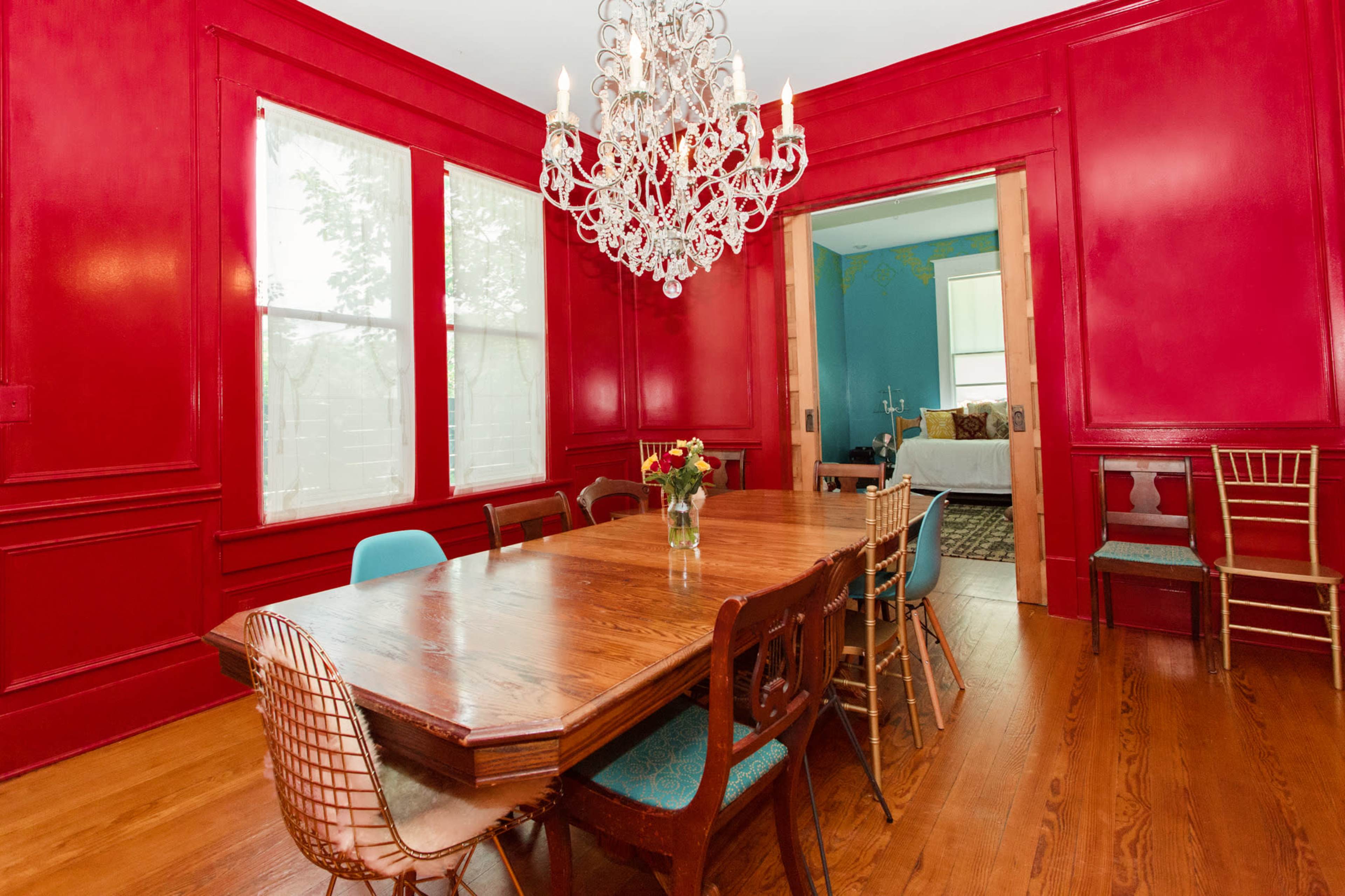 The dining room features a large wooden table surrounded by various chairs, with a crystal chandelier hanging from the ceiling and bright red walls.