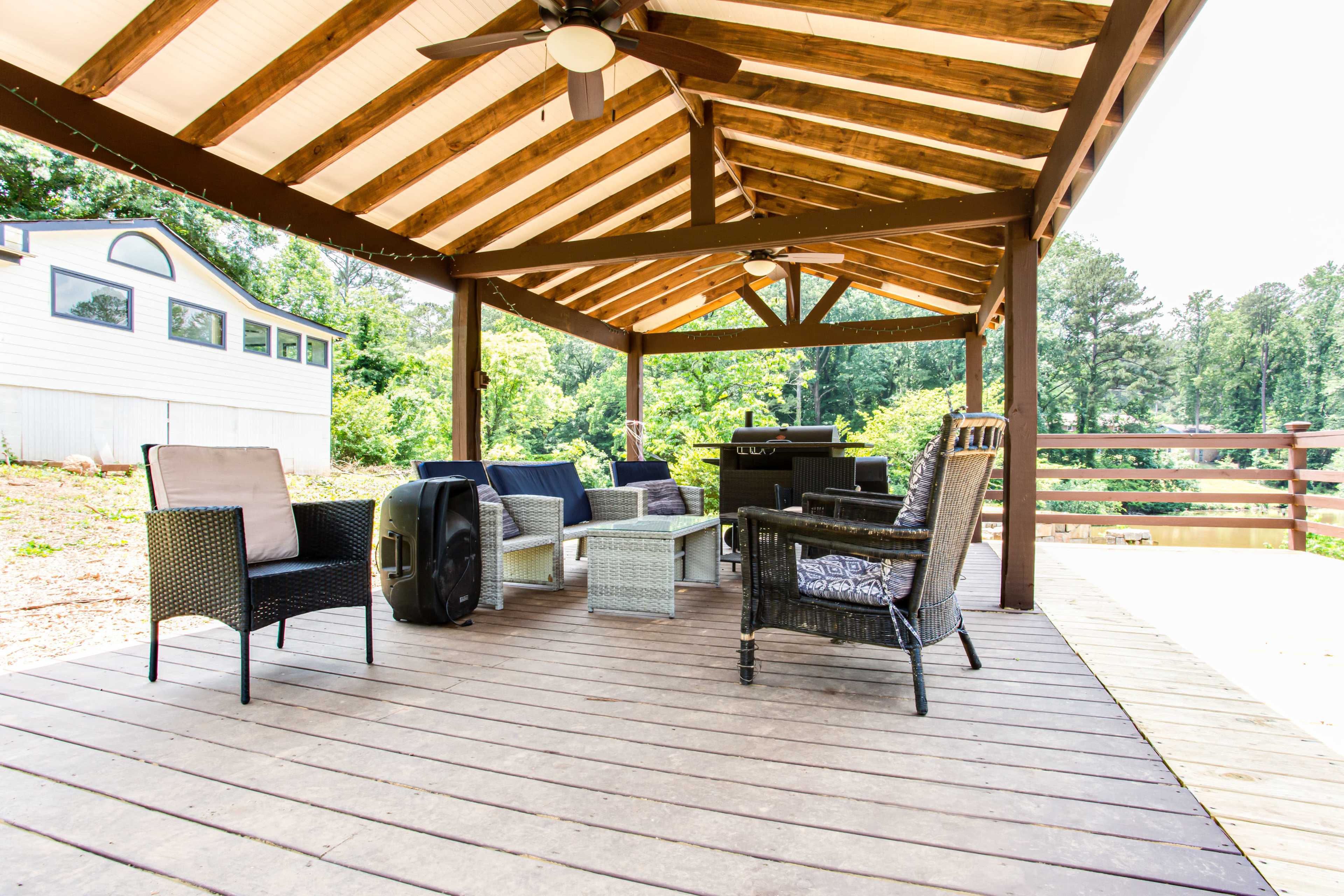 The image shows a covered outdoor seating area with a wooden roof, featuring several chairs arranged around a table and a grill.