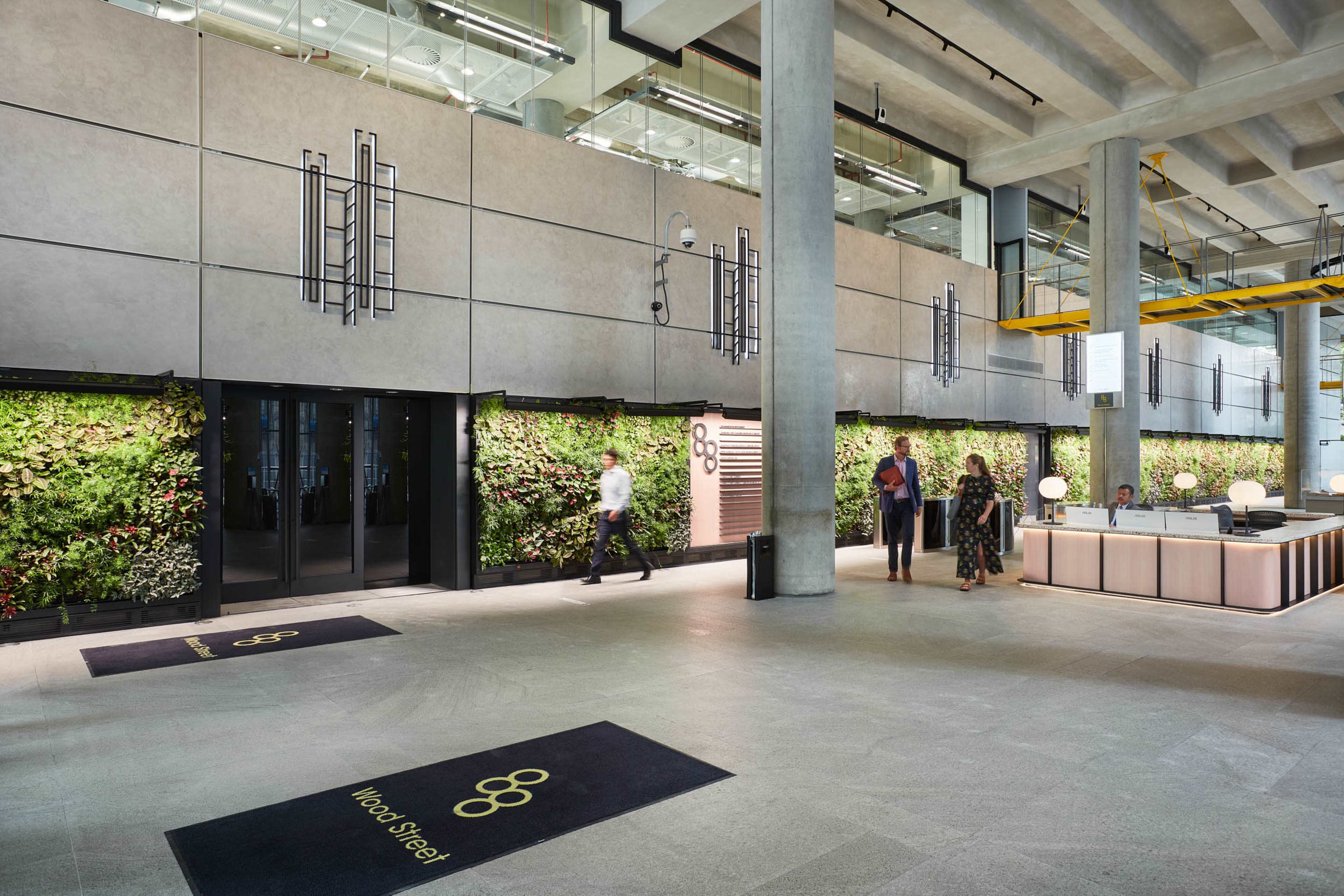 The image shows a modern office lobby featuring green wall installations, concrete columns, and a reception desk with visitors interacting in the space.