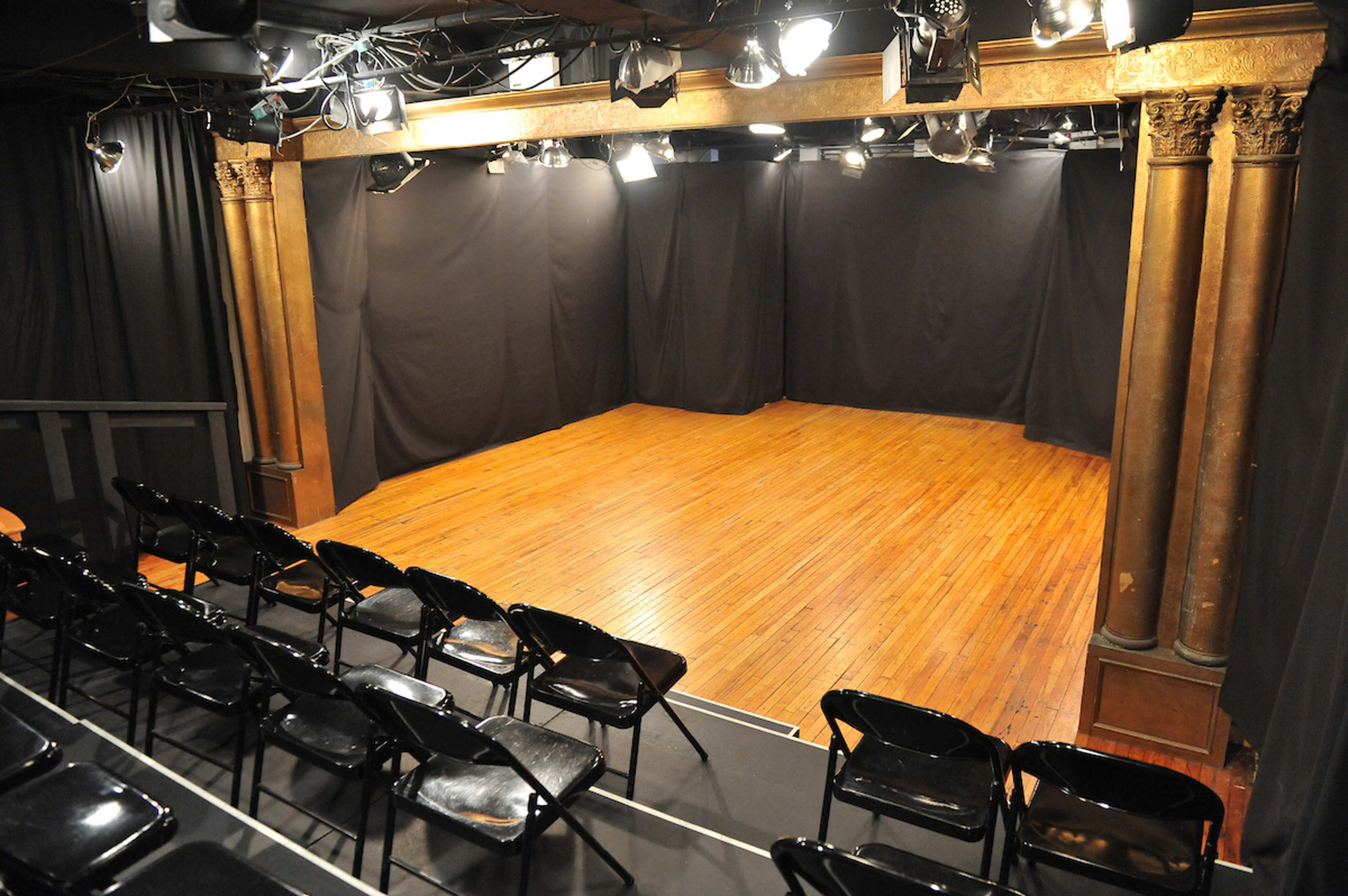 The image shows a small theater stage with wooden flooring and black drapes, accompanied by rows of black chairs facing the stage.
