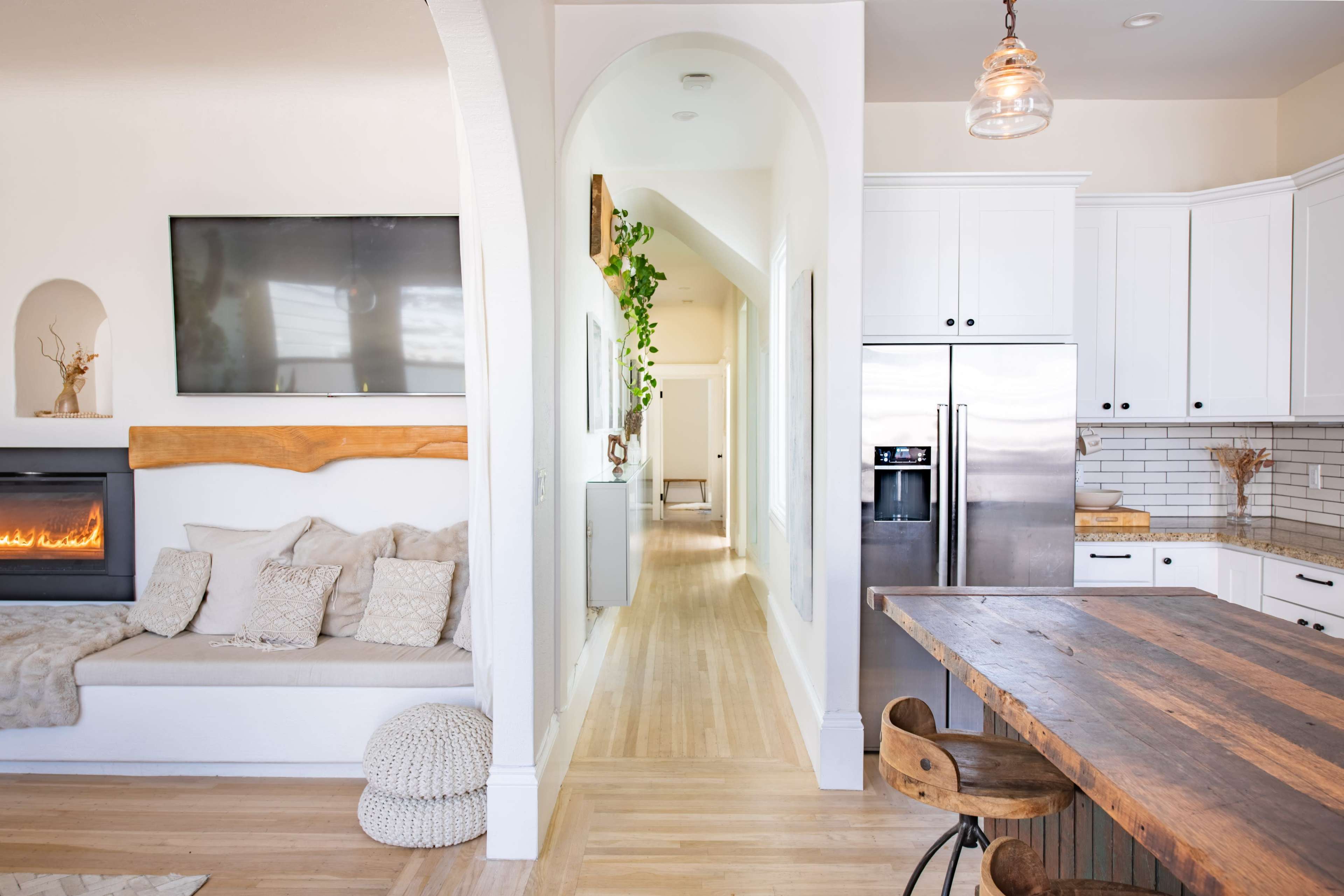 The image shows a modern living space with a cozy seating area beside a wall-mounted TV, an open kitchen featuring stainless steel appliances, and a light-filled hallway leading to another room.