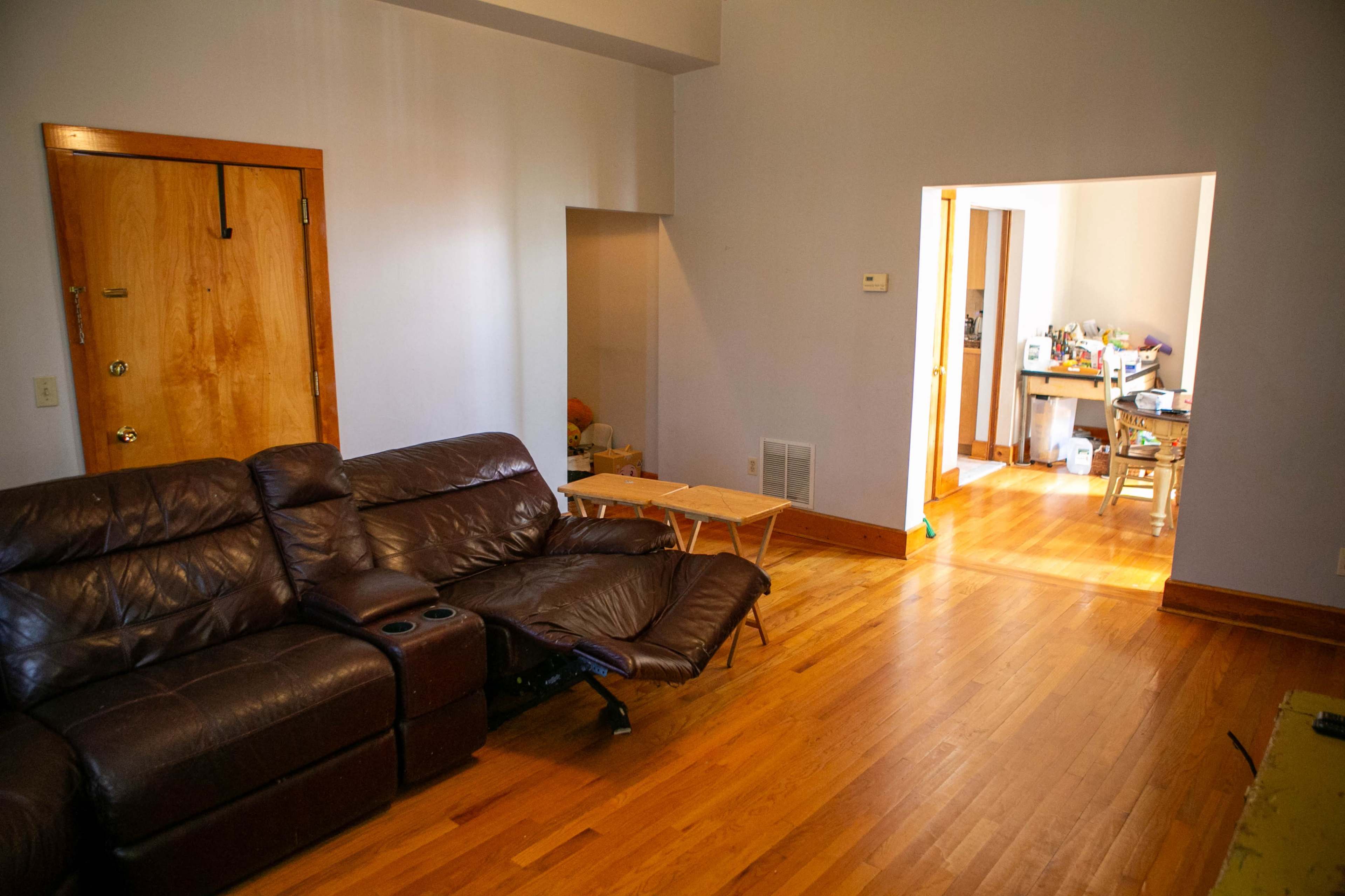 The image shows a spacious living room with a brown leather couch, a small folding table, and an adjoining doorway leading to a well-lit area with wooden floors and a cluttered table.