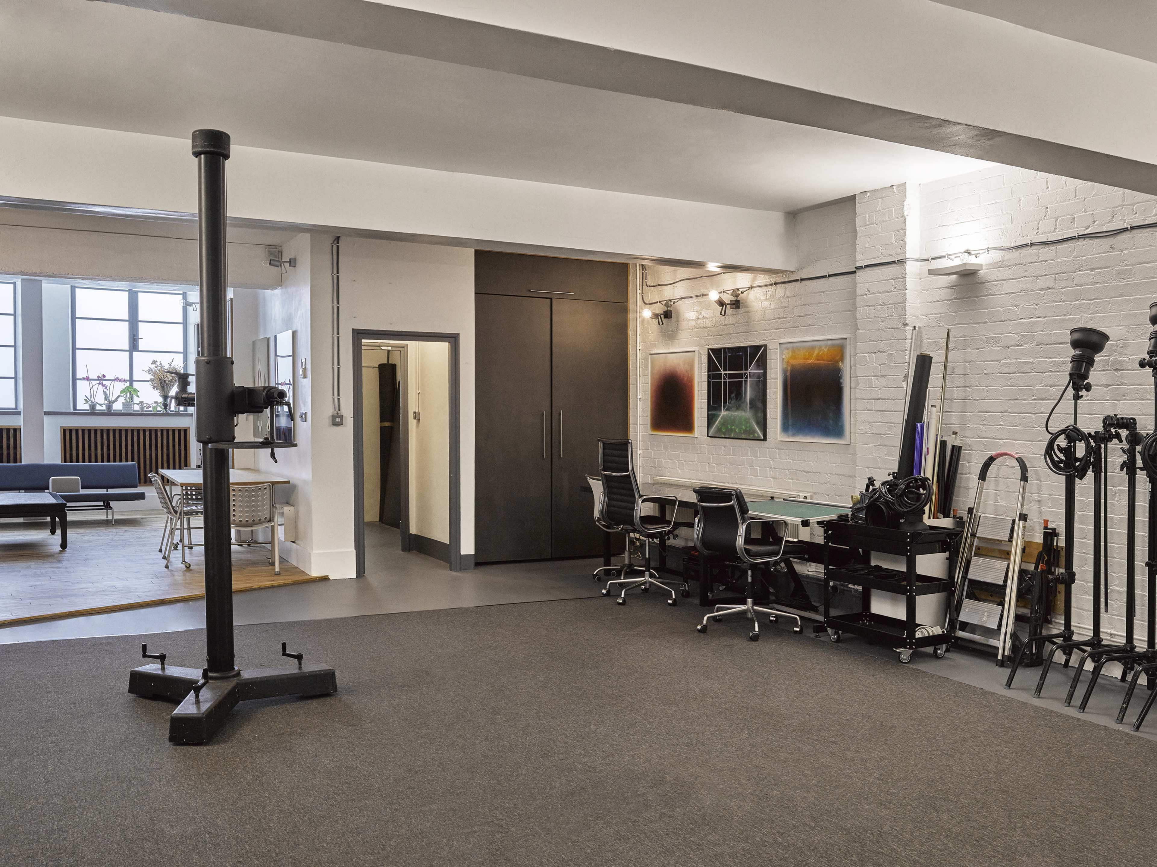 The image shows an open studio space with a desk area featuring black chairs, a large wall-mounted storage unit, and various equipment stored on carts along one side.