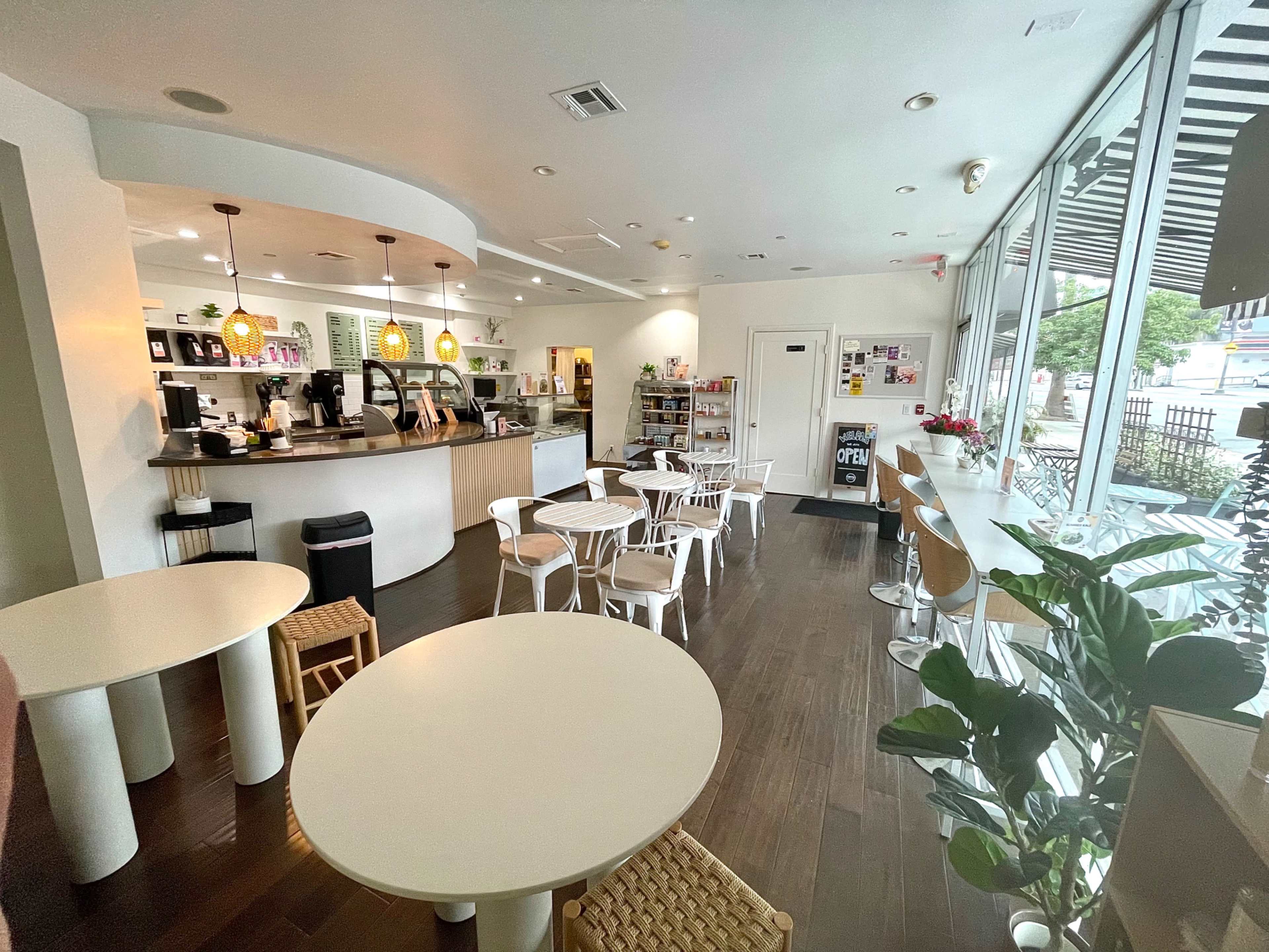 The image shows a bright café interior featuring tables and chairs, a counter with coffee equipment, and a display of products by the window.