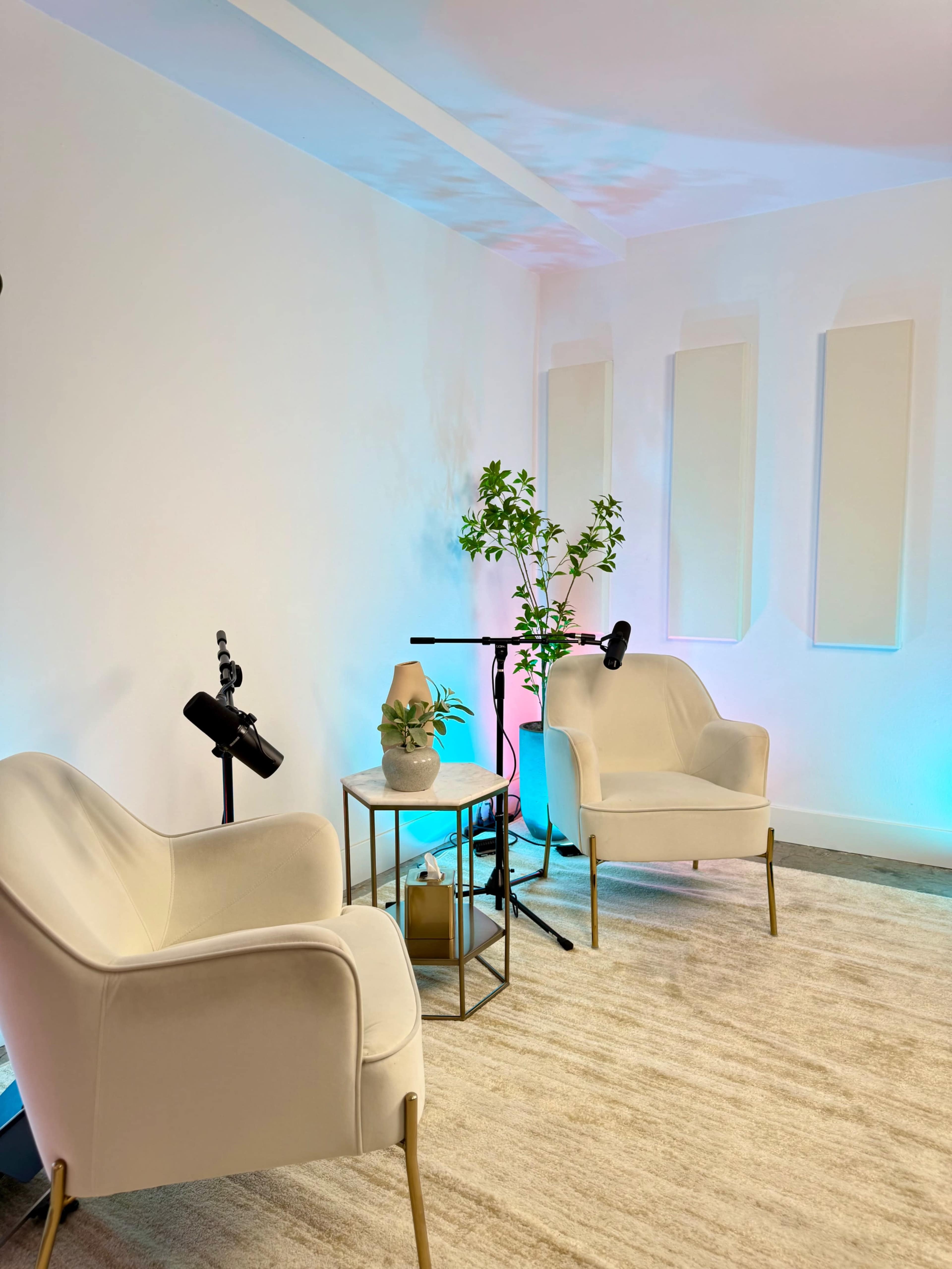 The image shows a minimalist room featuring two white armchairs, a small metal side table, and plants, illuminated by soft, colorful lighting.